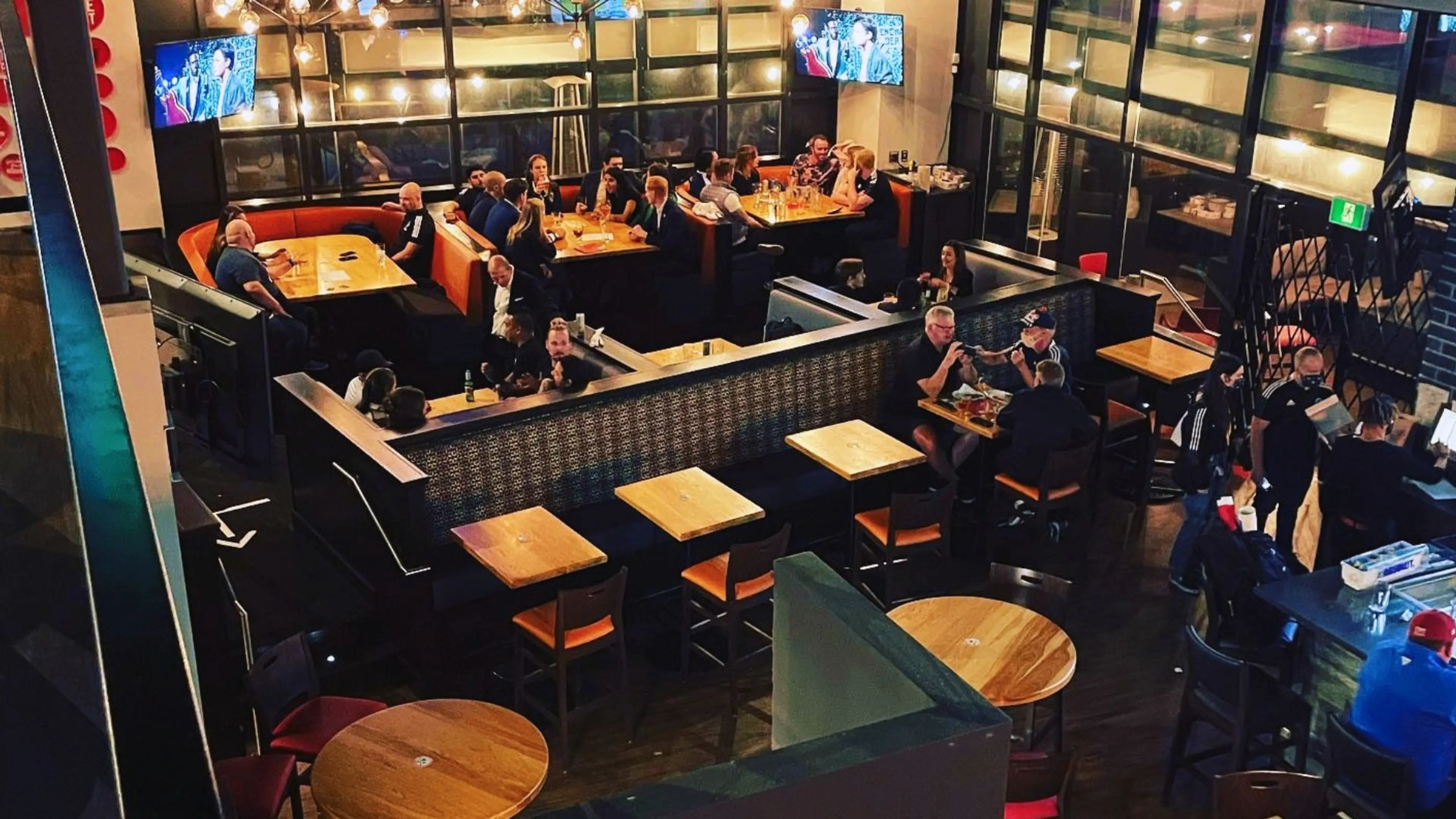 Interior of a busy restaurant with people sitting at tables and booths, two TVs mounted on the walls.