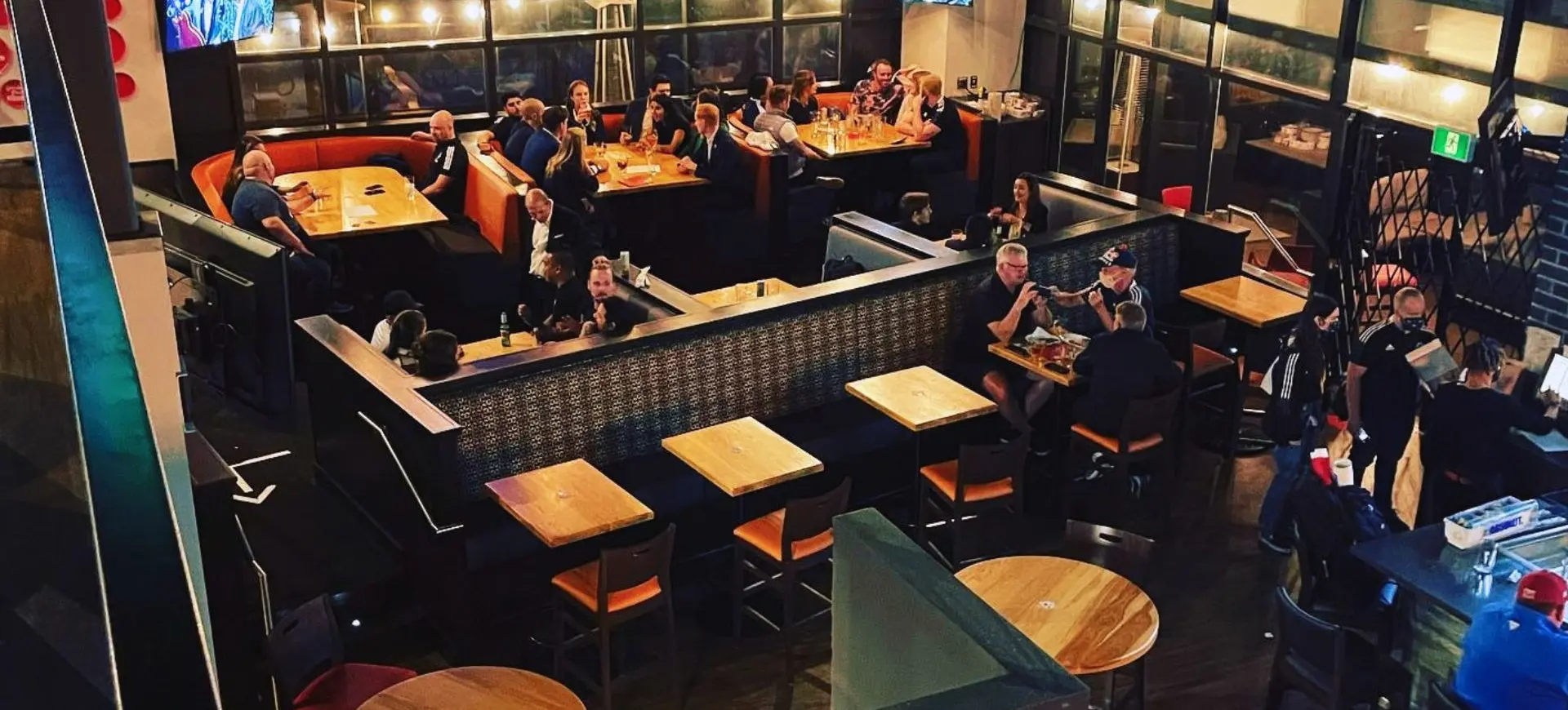 Interior of a busy restaurant with people sitting at tables and booths, two TVs mounted on the walls.