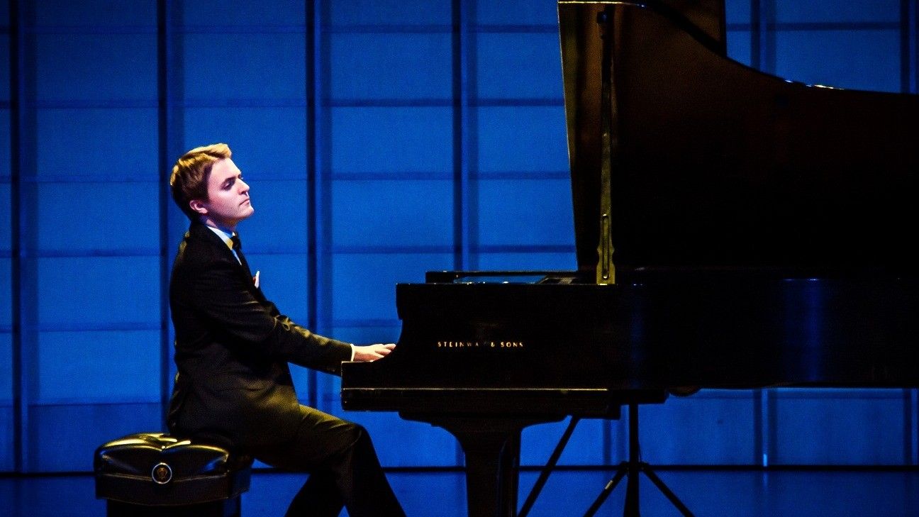 Person in a black suit playing a grand piano on a stage with a blue background.