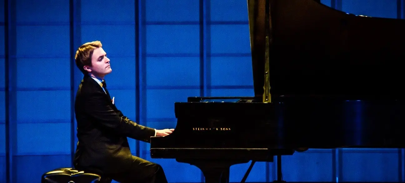 Person in a black suit playing a grand piano on a stage with a blue background.