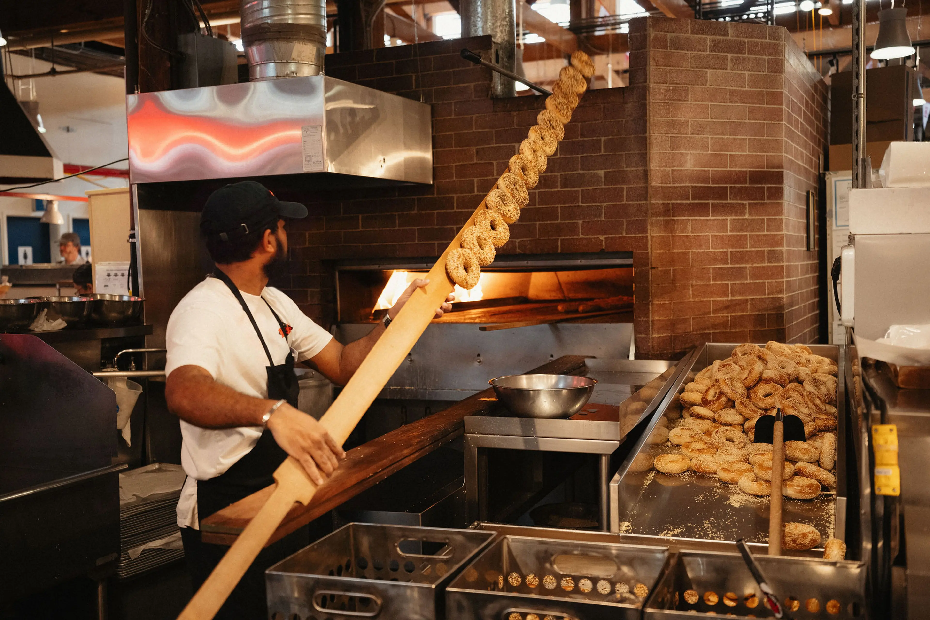 A person using a long wooden paddle to place or remove bagels from a brick oven in a bakery kitchen.