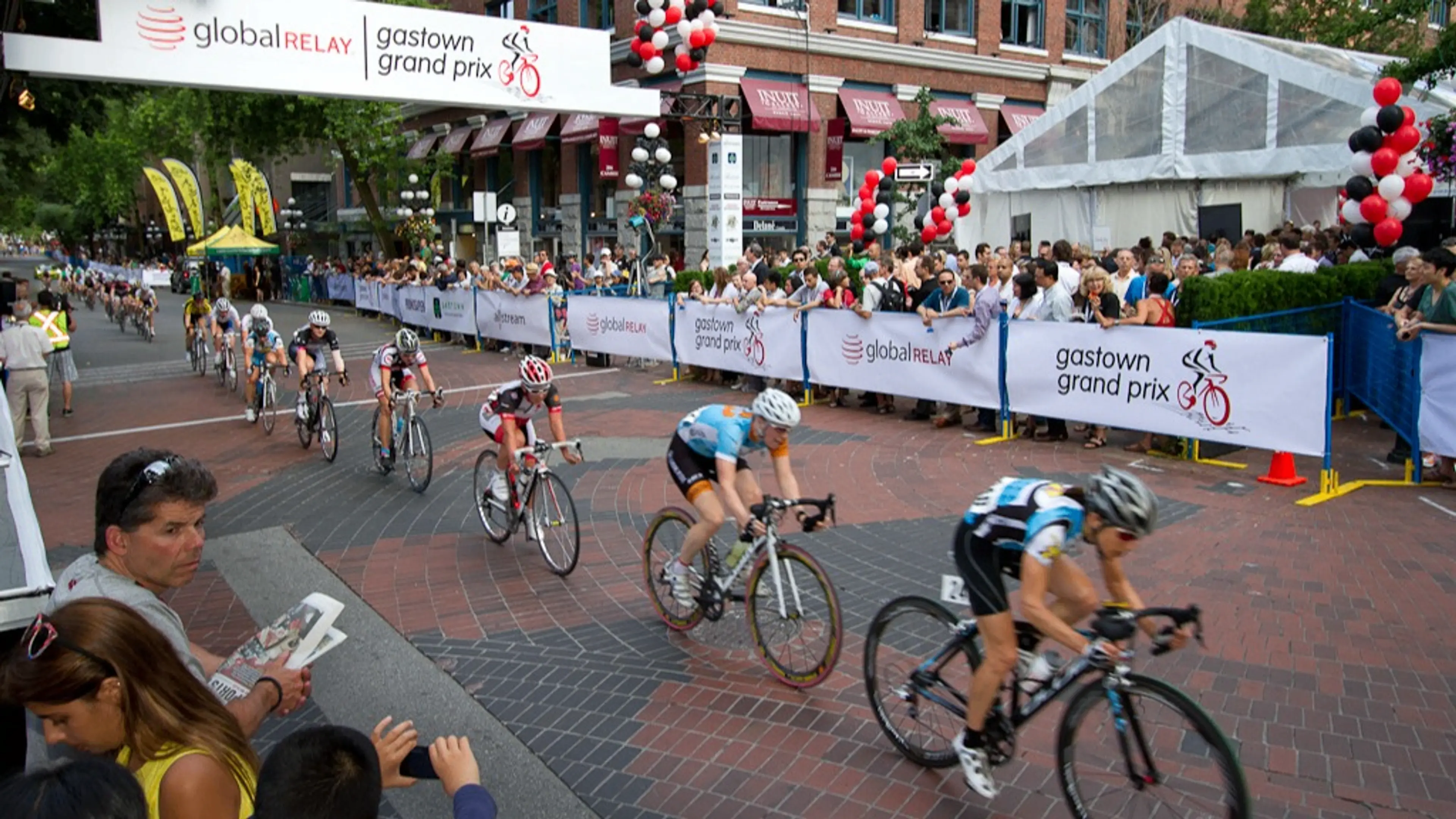 Cyclists racing on a brick-paved street during the Gastown Grand Prix with spectators behind barriers and event banners overhead.