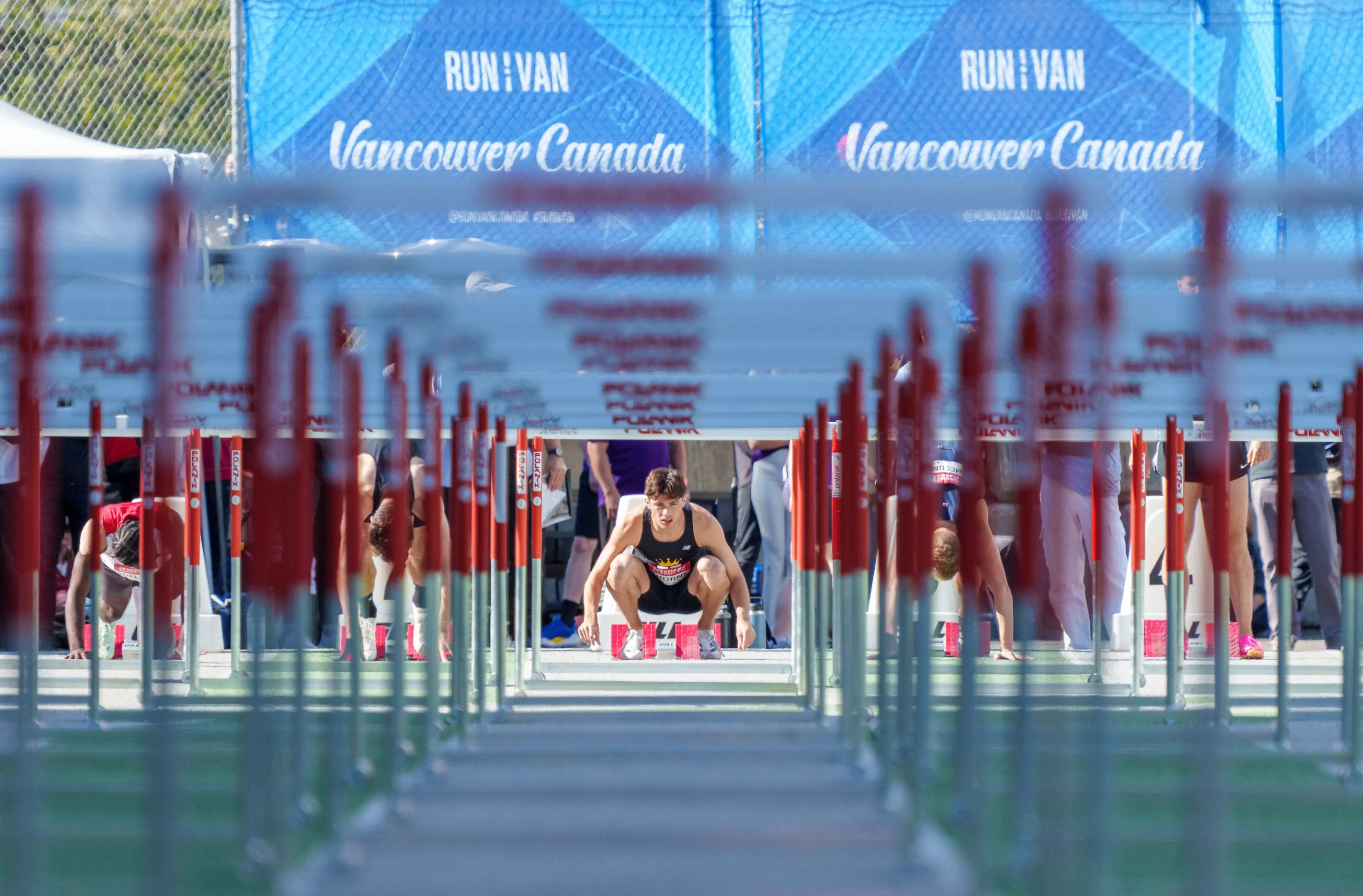 Athletes crouched at the start line of a hurdle race on a track with a blue banner reading 'Run Vancouver Canada' in the background.