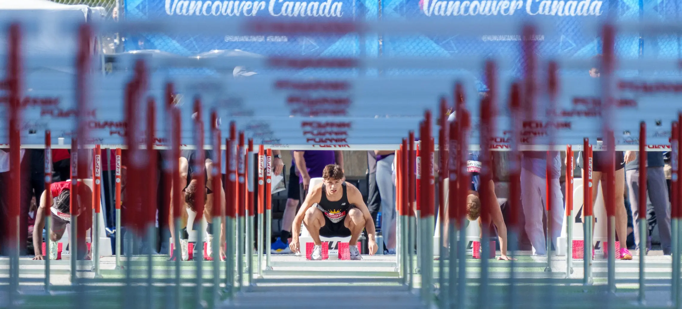 Athletes crouched at the start line of a hurdle race on a track with a blue banner reading 'Run Vancouver Canada' in the background.