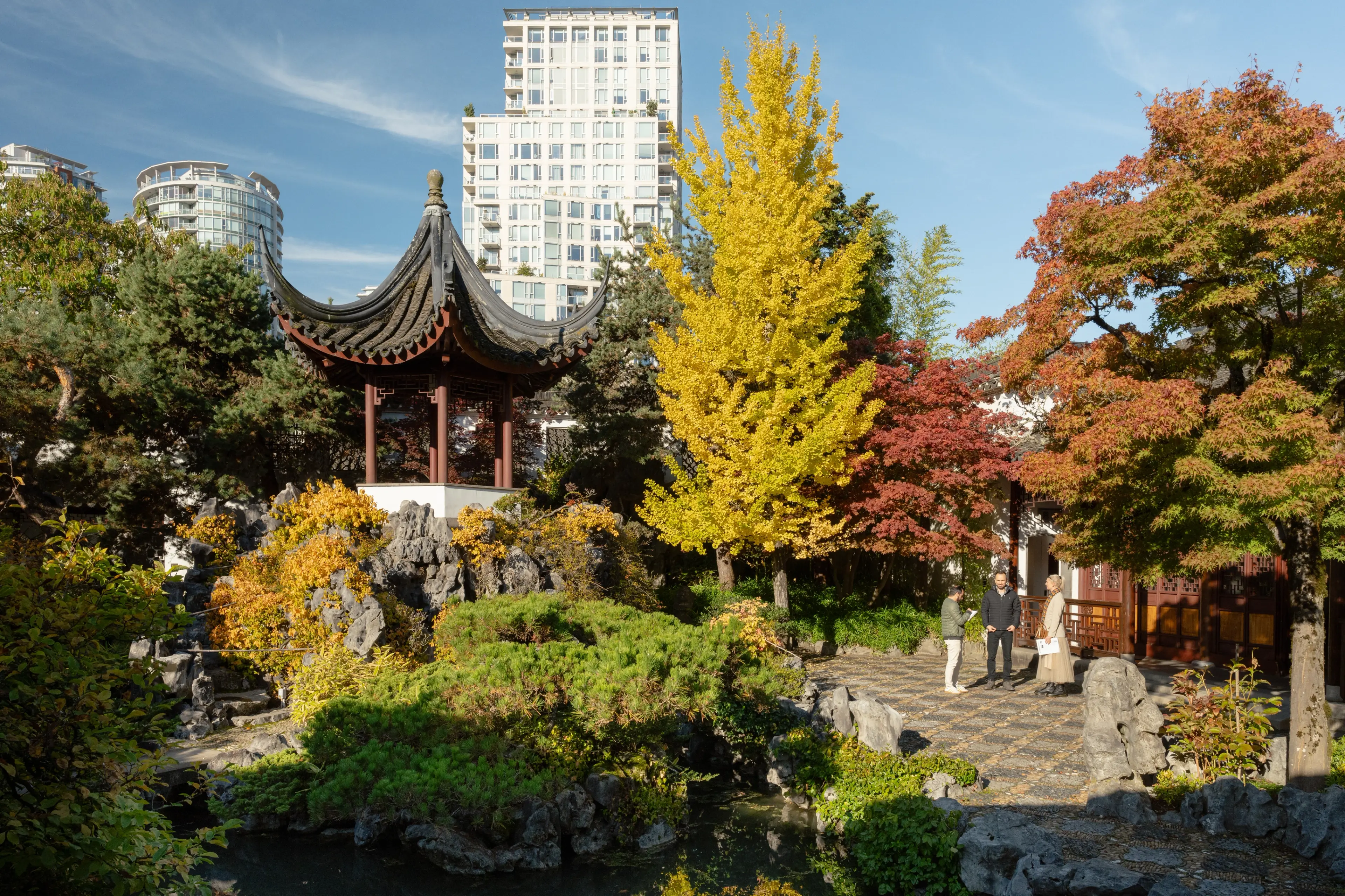 Three people standing and talking in a garden with autumn trees, rocks, a pond, and a traditional pagoda, with tall buildings in the background.