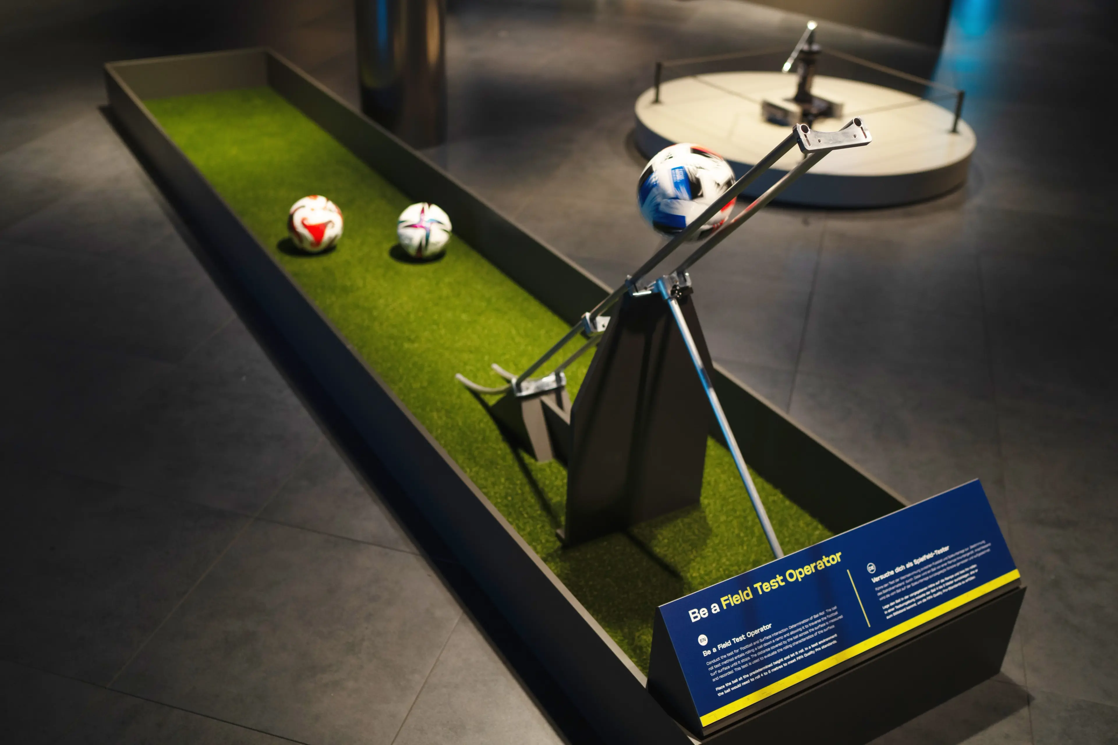 A mechanical device on a green turf platform with three soccer balls, one held by the device, and a sign titled 'Be a Field Test Operator'.