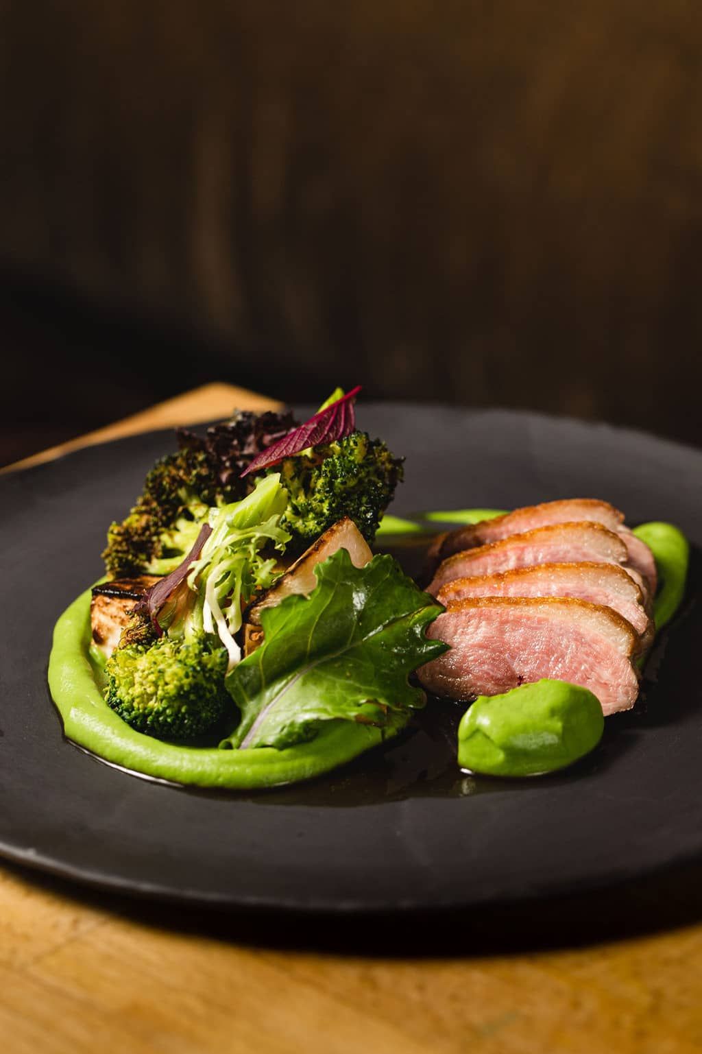 Sliced cooked meat with green vegetable puree, broccoli, and leafy greens on a black plate.