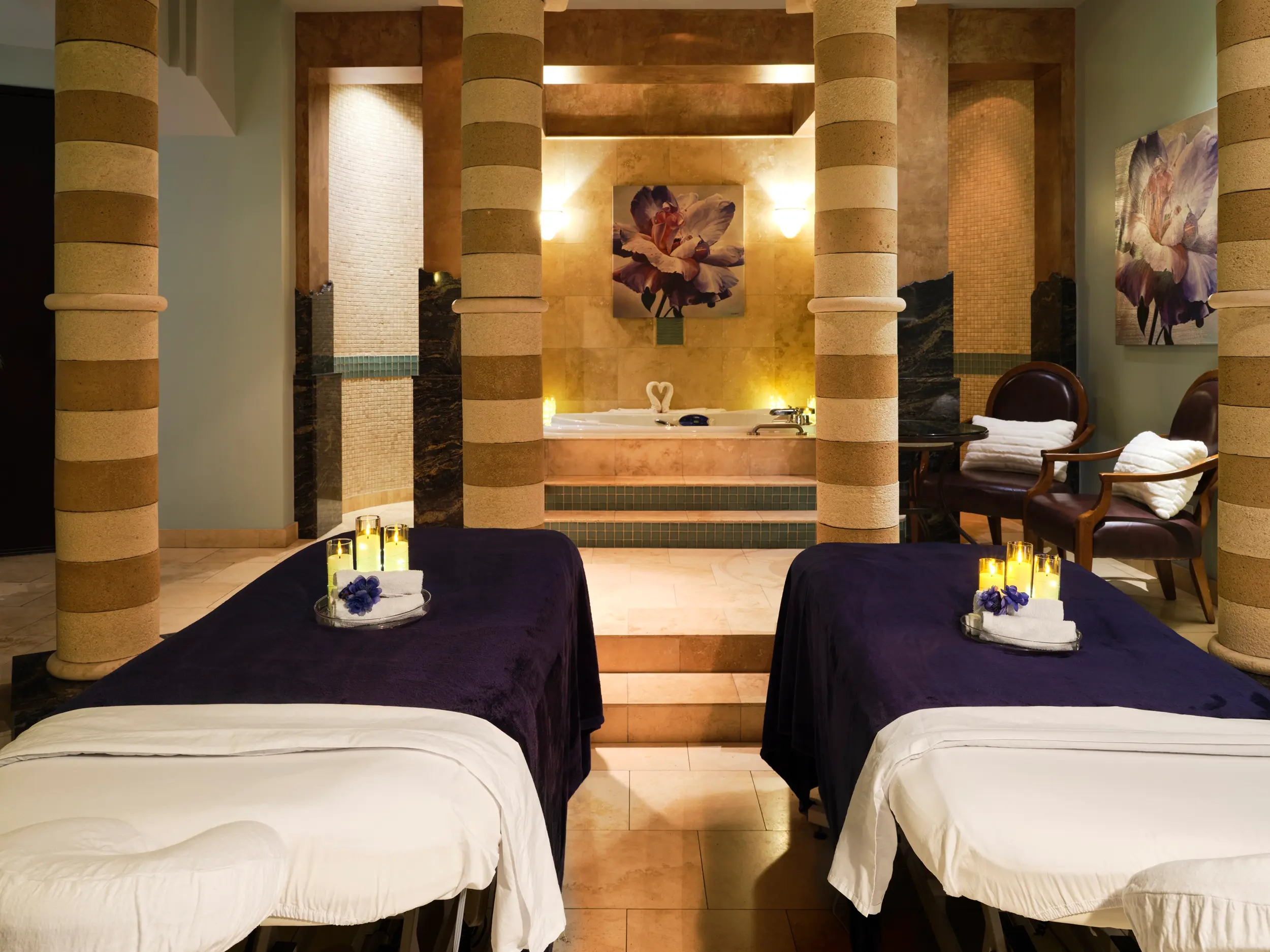 Two massage tables with dark covers and white sheets in a spa room with candles, towels, and flower decorations, and a bathtub in the background.