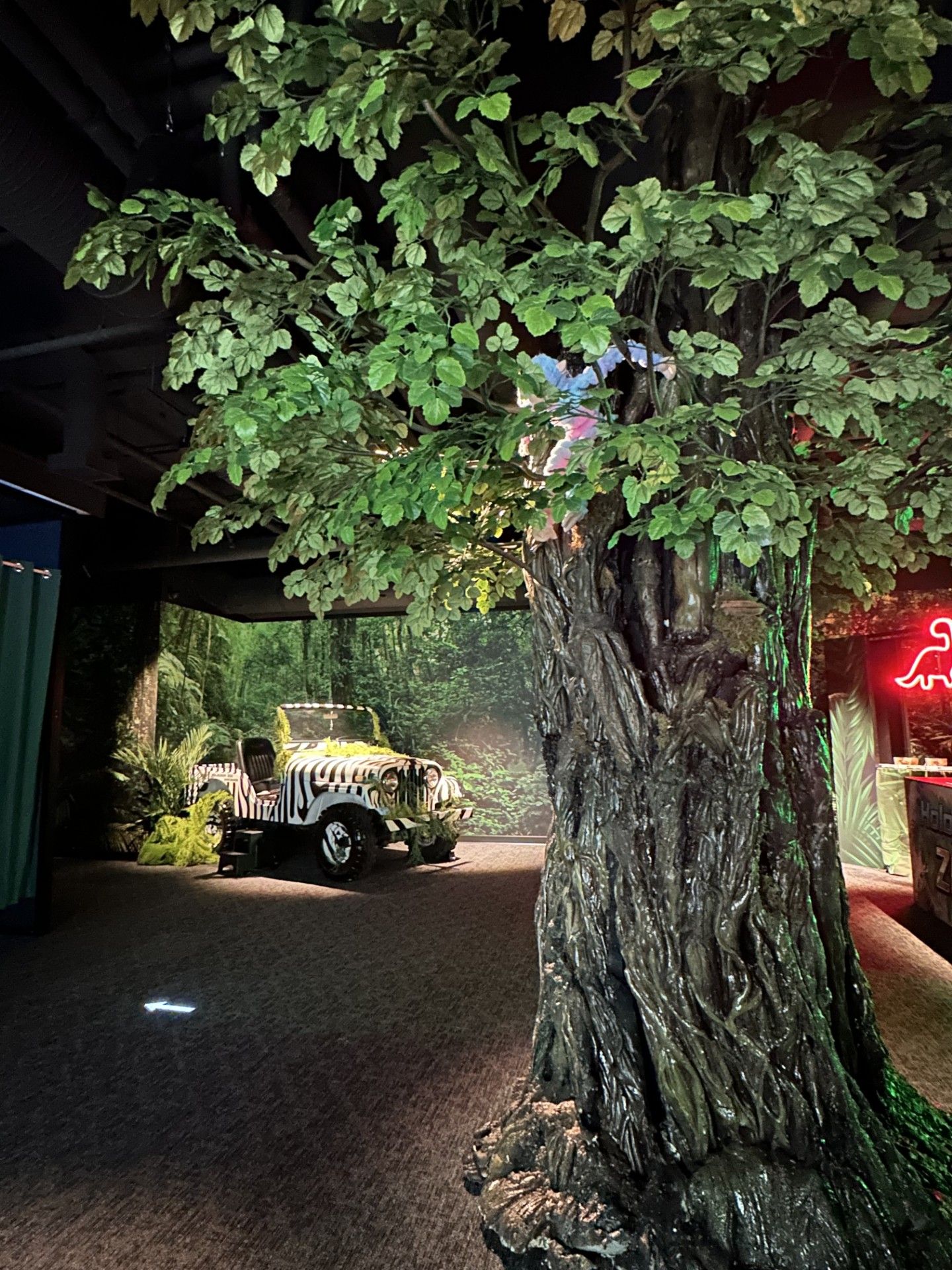 Large artificial tree trunk with green leaves indoors, zebra-striped jeep and jungle-themed decor in background.