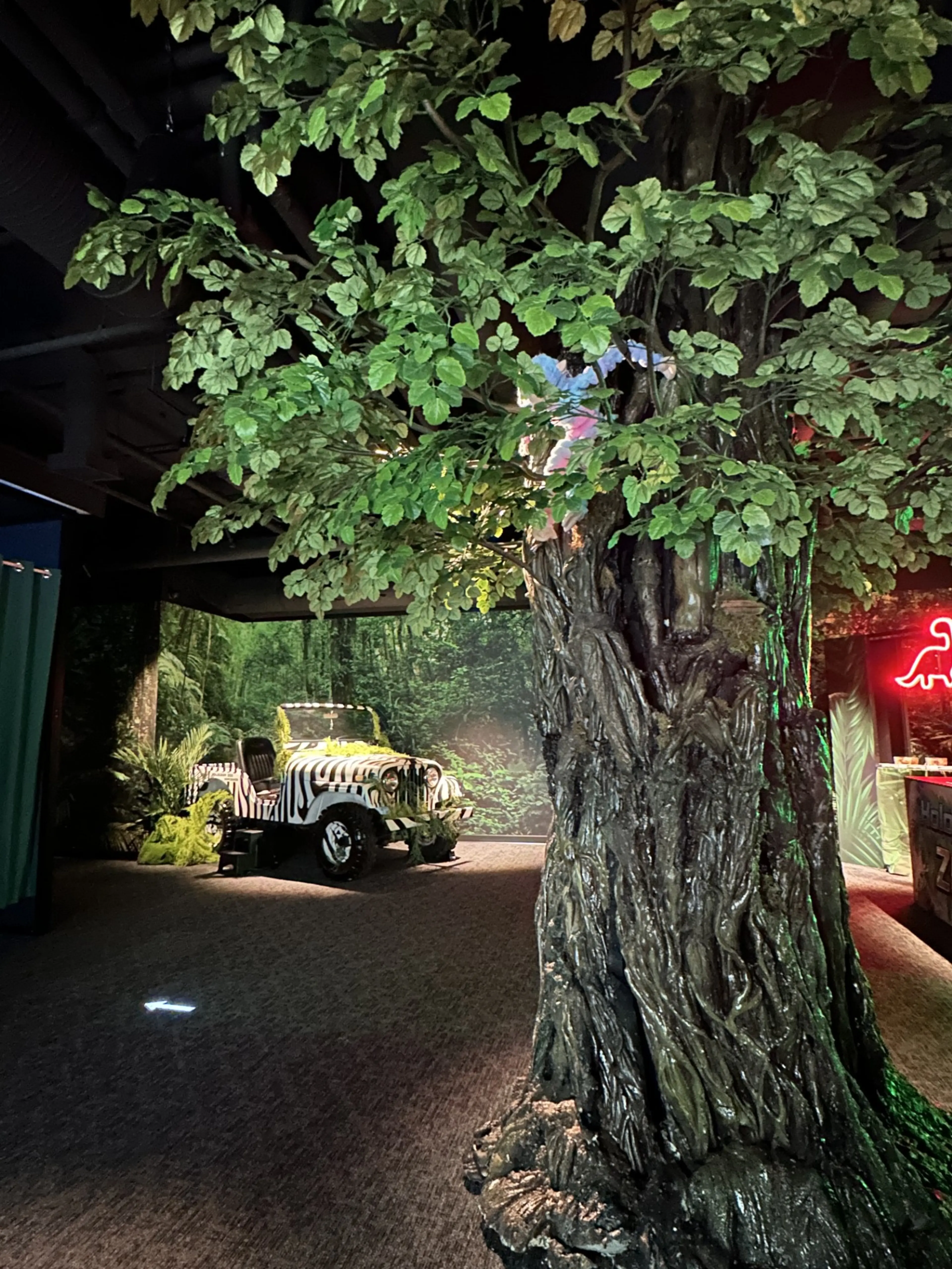 Large artificial tree trunk with green leaves indoors, zebra-striped jeep and jungle-themed decor in background.