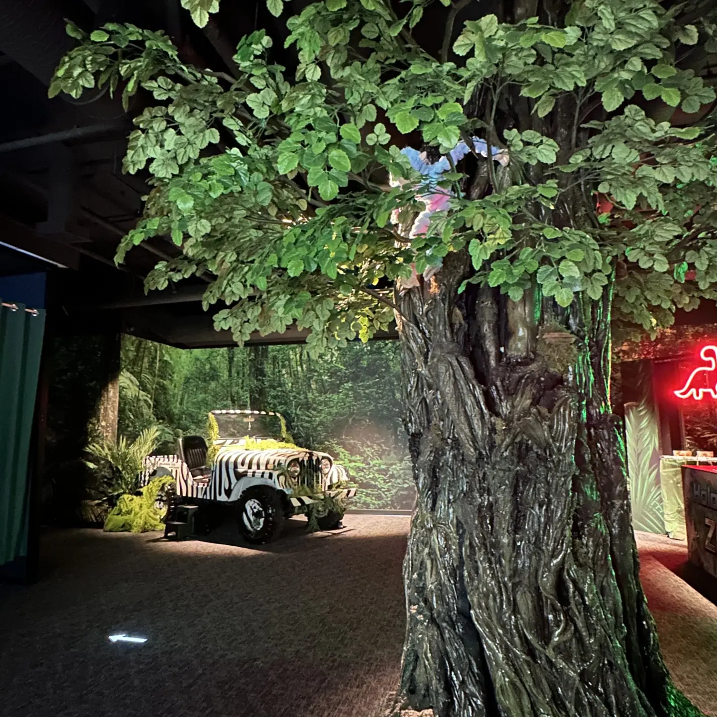 Large artificial tree trunk with green leaves indoors, zebra-striped jeep and jungle-themed decor in background.