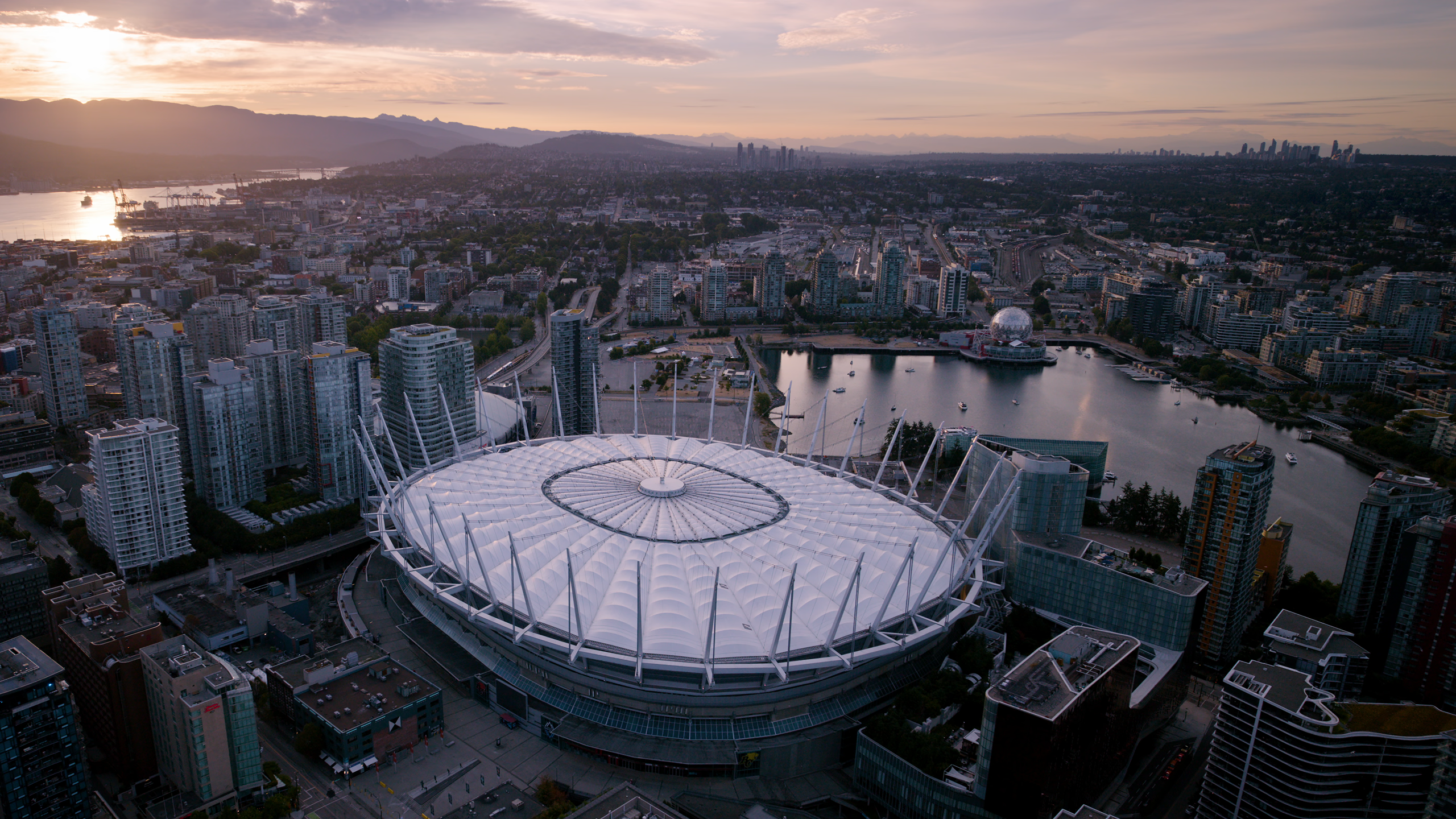 Aerial view of a large stadium with a white roof surrounded by city buildings and a body of water at sunset.