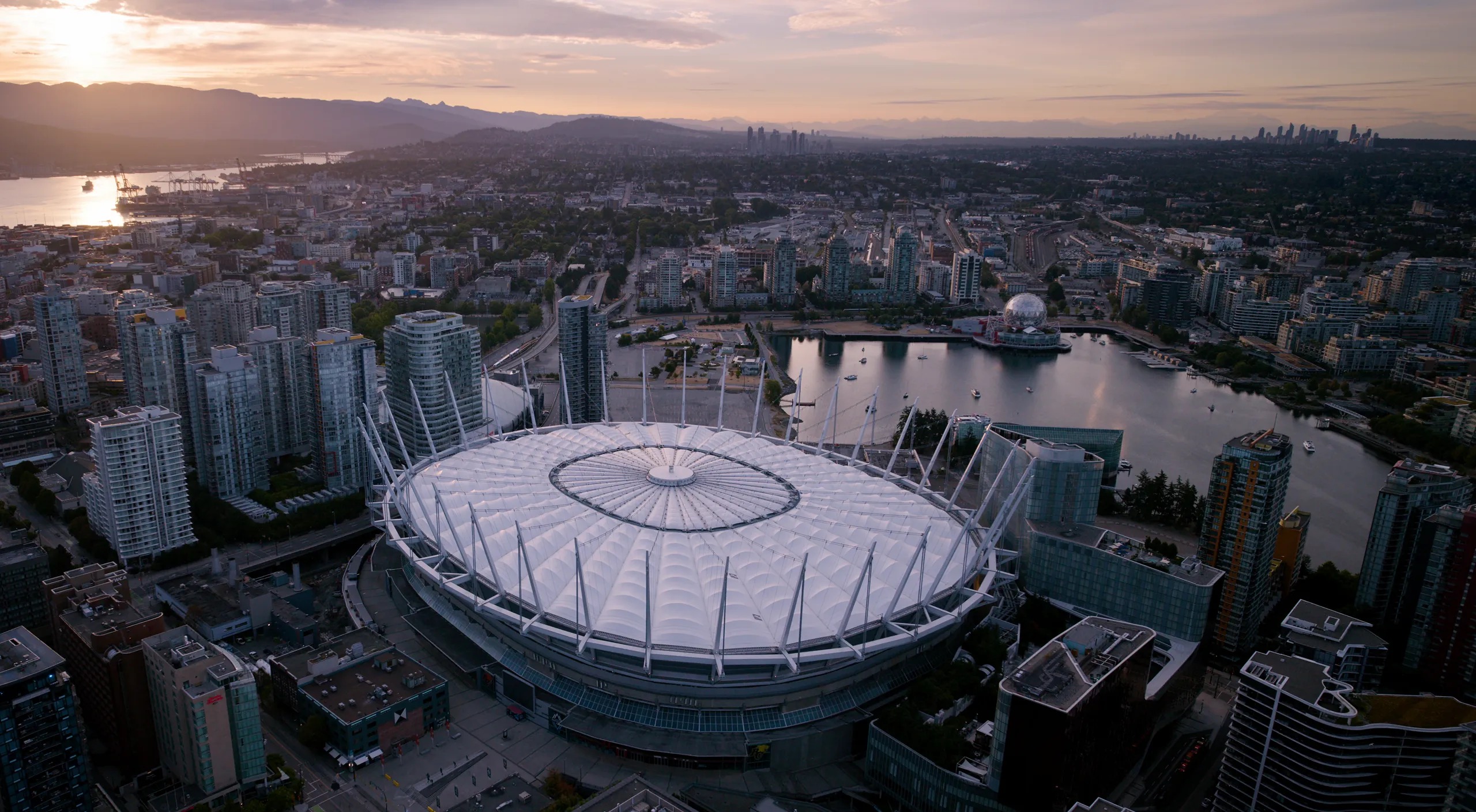 Aerial view of a large stadium with a white roof surrounded by city buildings and water at sunset.