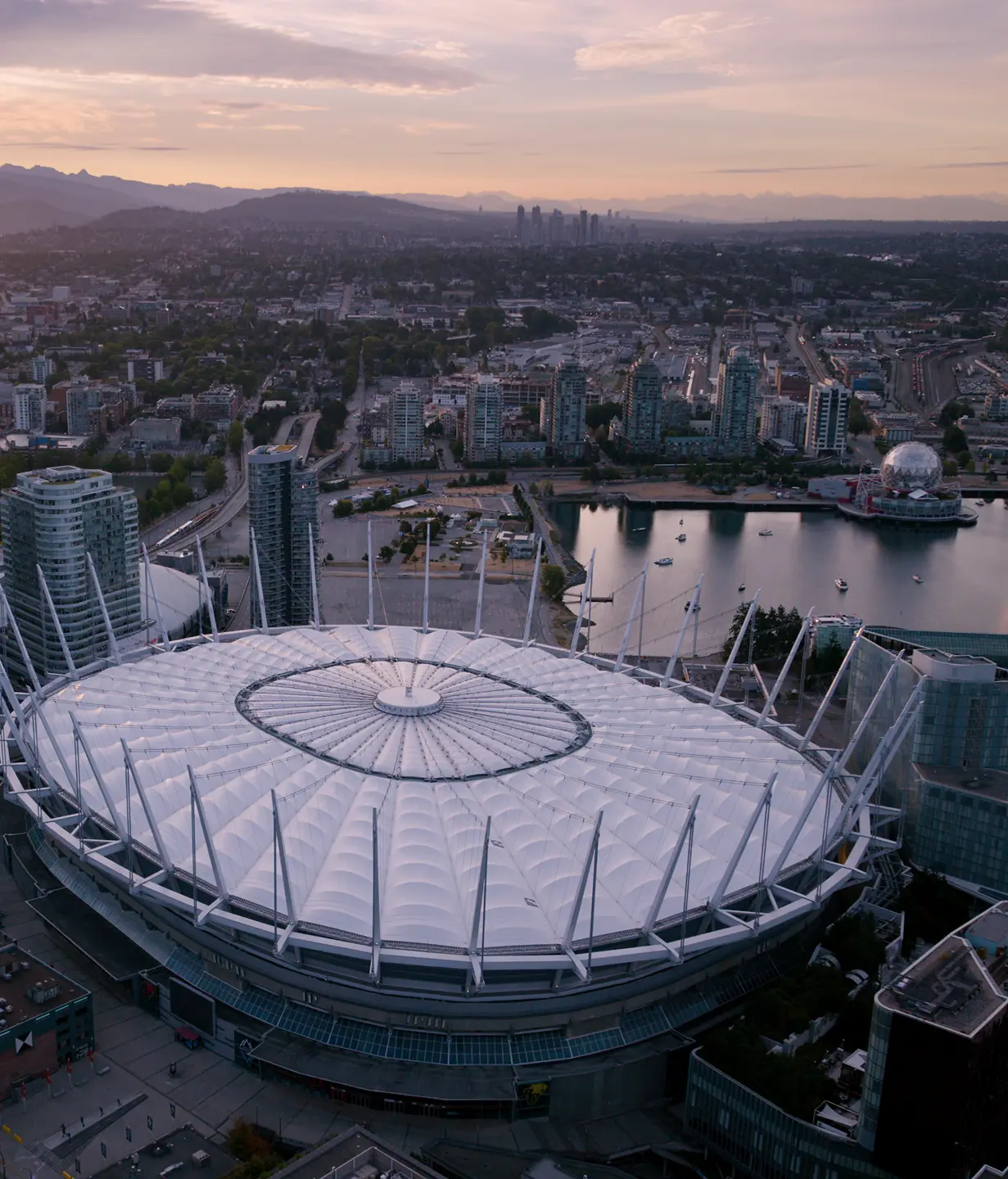 Aerial view of a large stadium with a white roof surrounded by city buildings and a body of water at sunset.