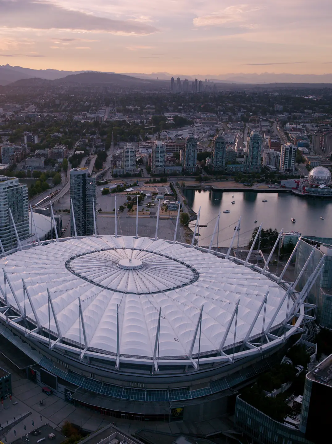 Aerial view of a large stadium with a white roof surrounded by city buildings and a body of water at sunset.