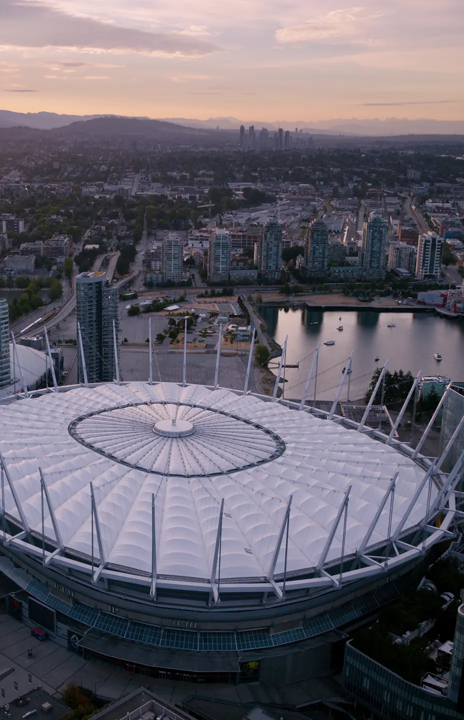 Aerial view of a large stadium with a white roof surrounded by city buildings and a body of water at sunset.