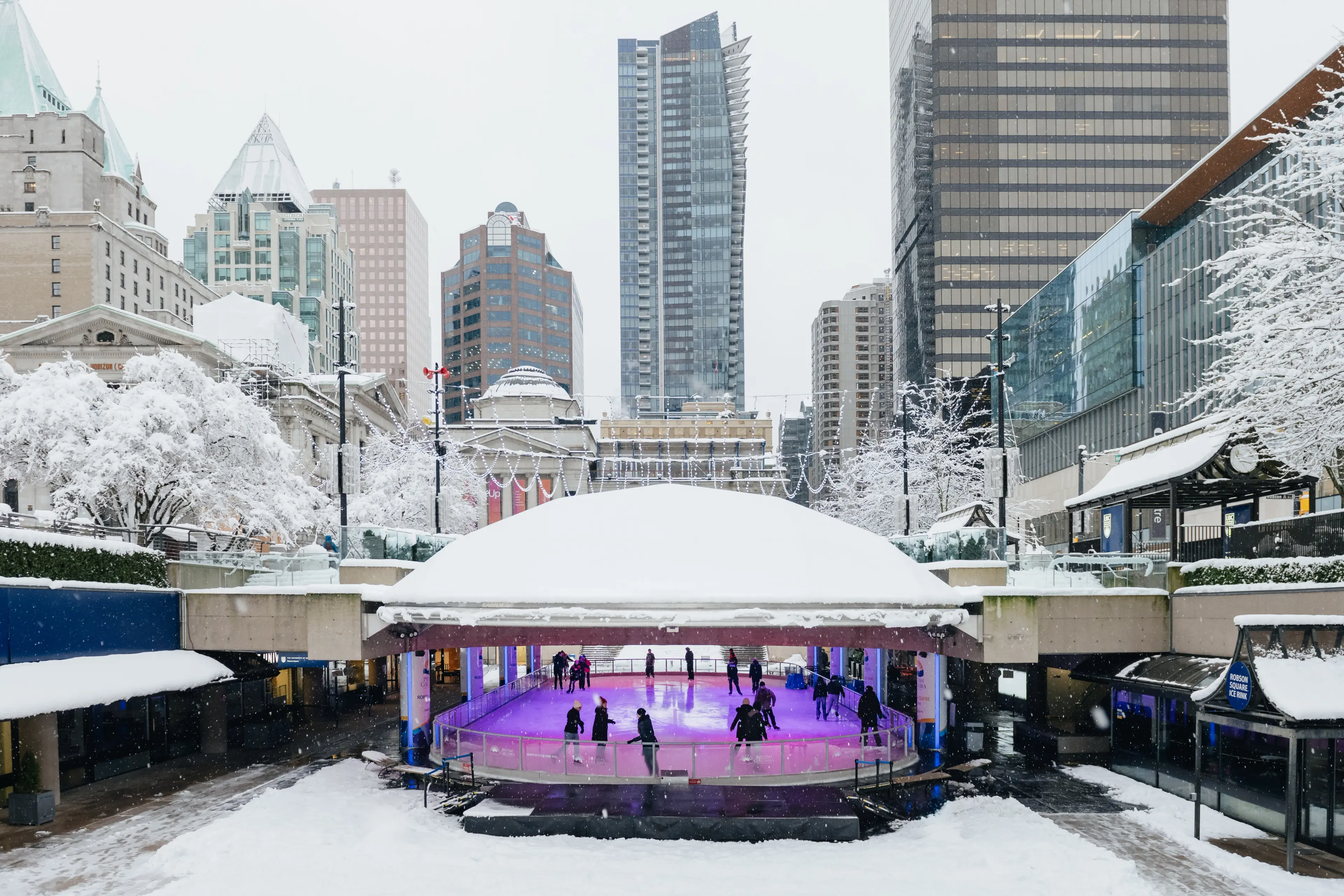 Skaters under the dome at the Robson Square Ice Rink. The surrounding city is covered in snow. 