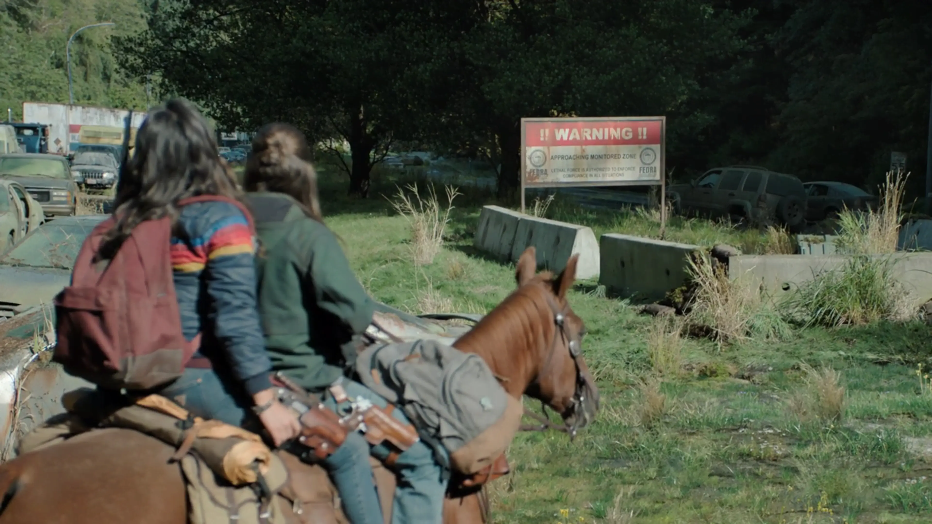 Two people riding a brown horse past a warning sign in a grassy area with abandoned cars and concrete barriers.