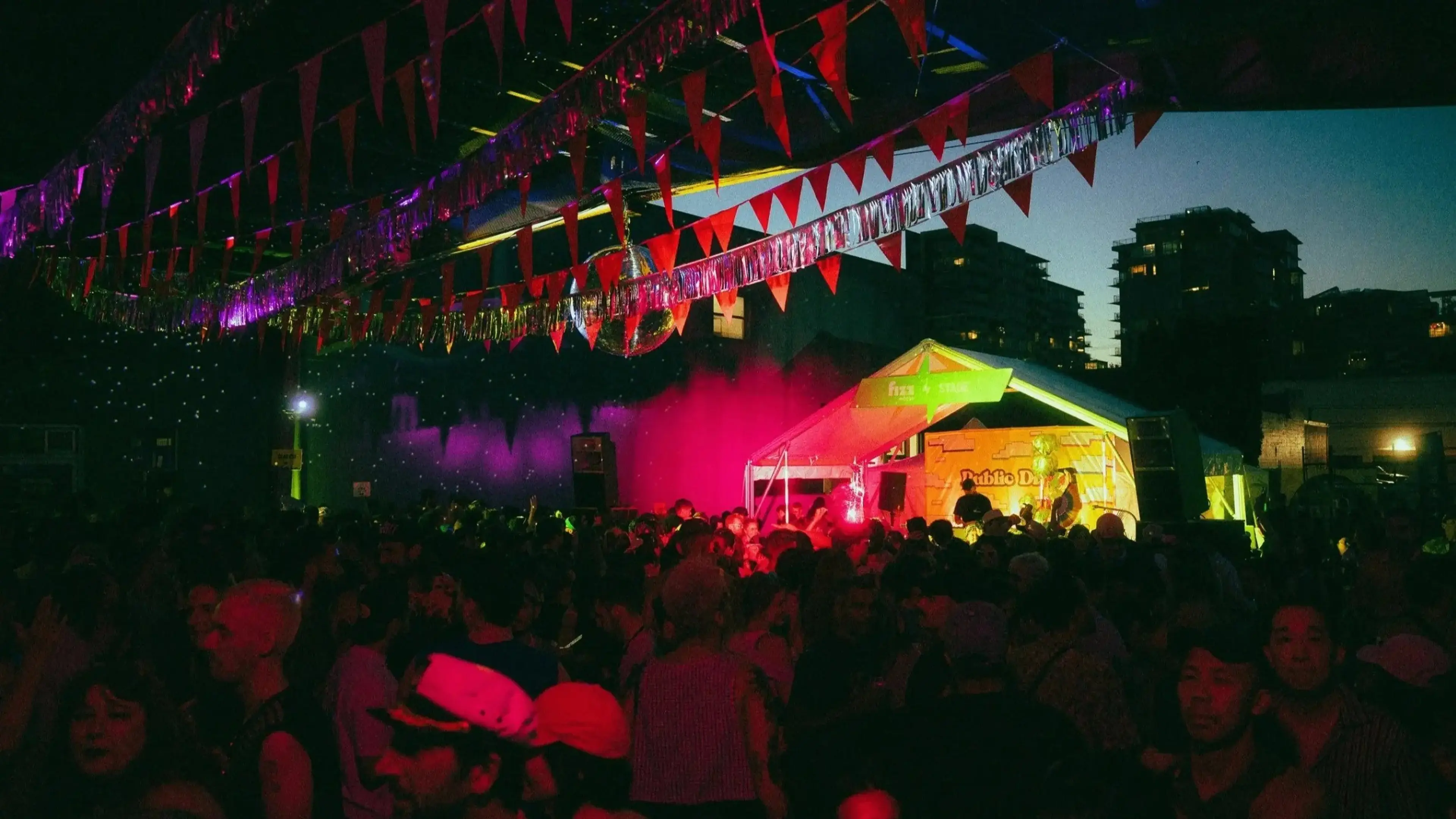Crowd gathered at an outdoor night event with colorful lights, a tent stage, and red triangular flags overhead.