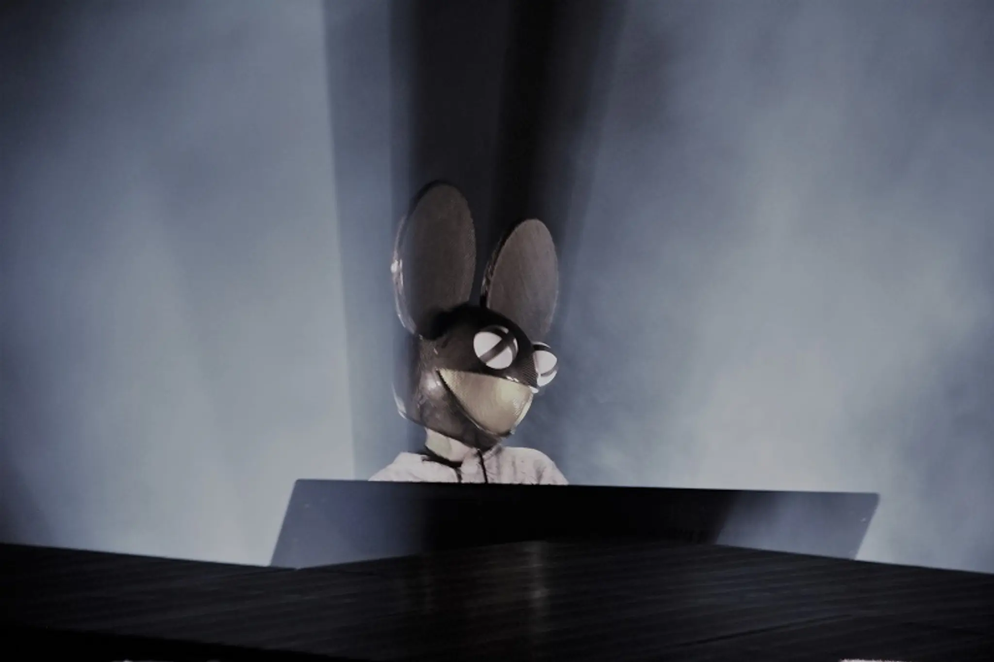 Person wearing a large mouse head mask with glasses, standing behind a DJ booth with blue lighting.
