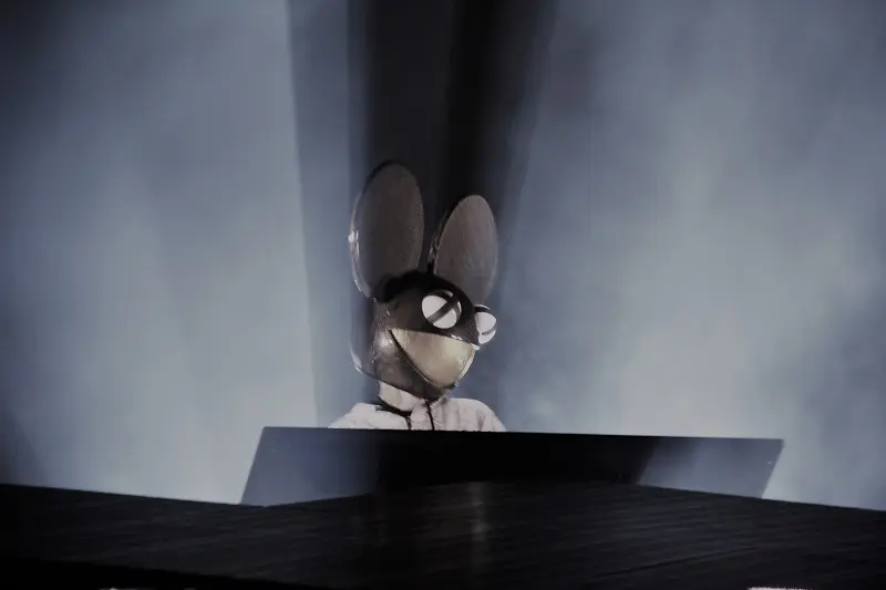 Person wearing a large mouse head mask with glasses, standing behind a DJ booth with blue lighting.