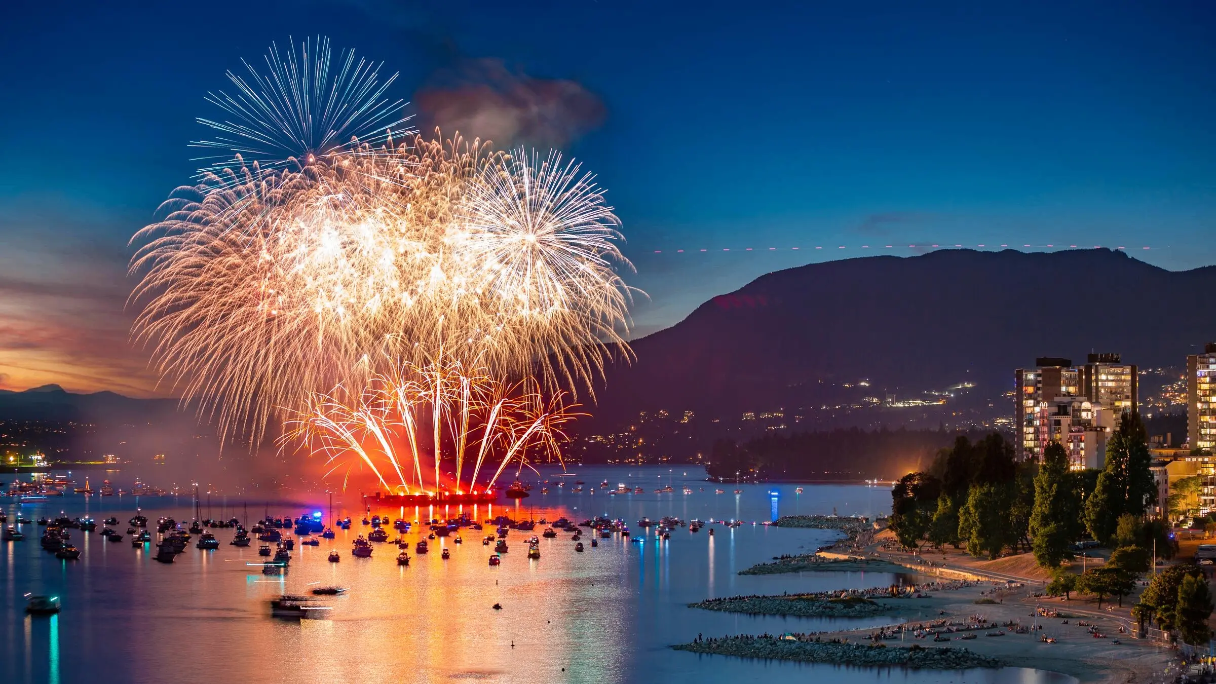 Fireworks exploding over water with boats watching, city buildings and mountains in the background at dusk.