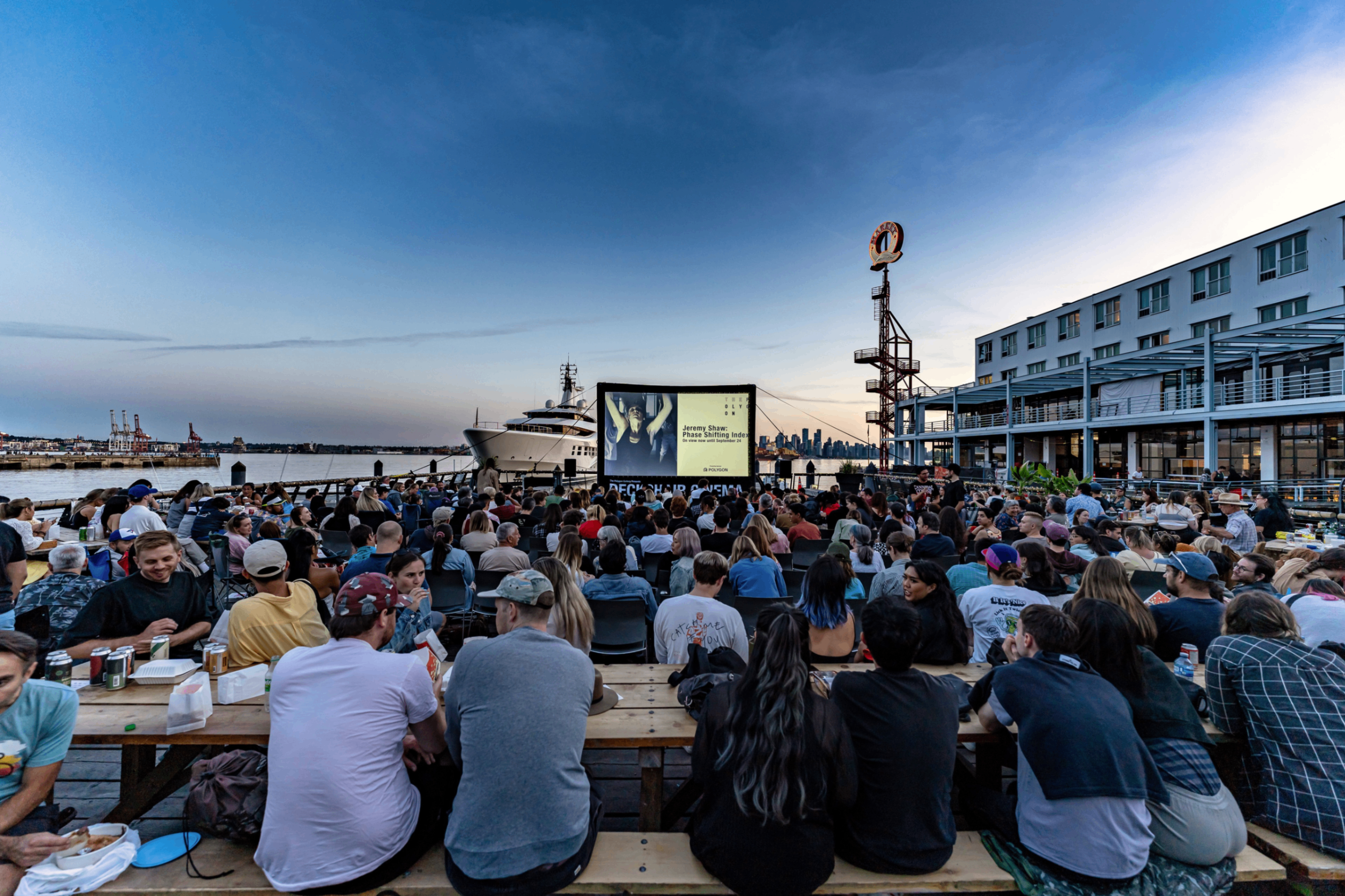Large crowd seated outdoors on benches watching a movie on a screen near a waterfront with a ship and city skyline in the background at dusk.