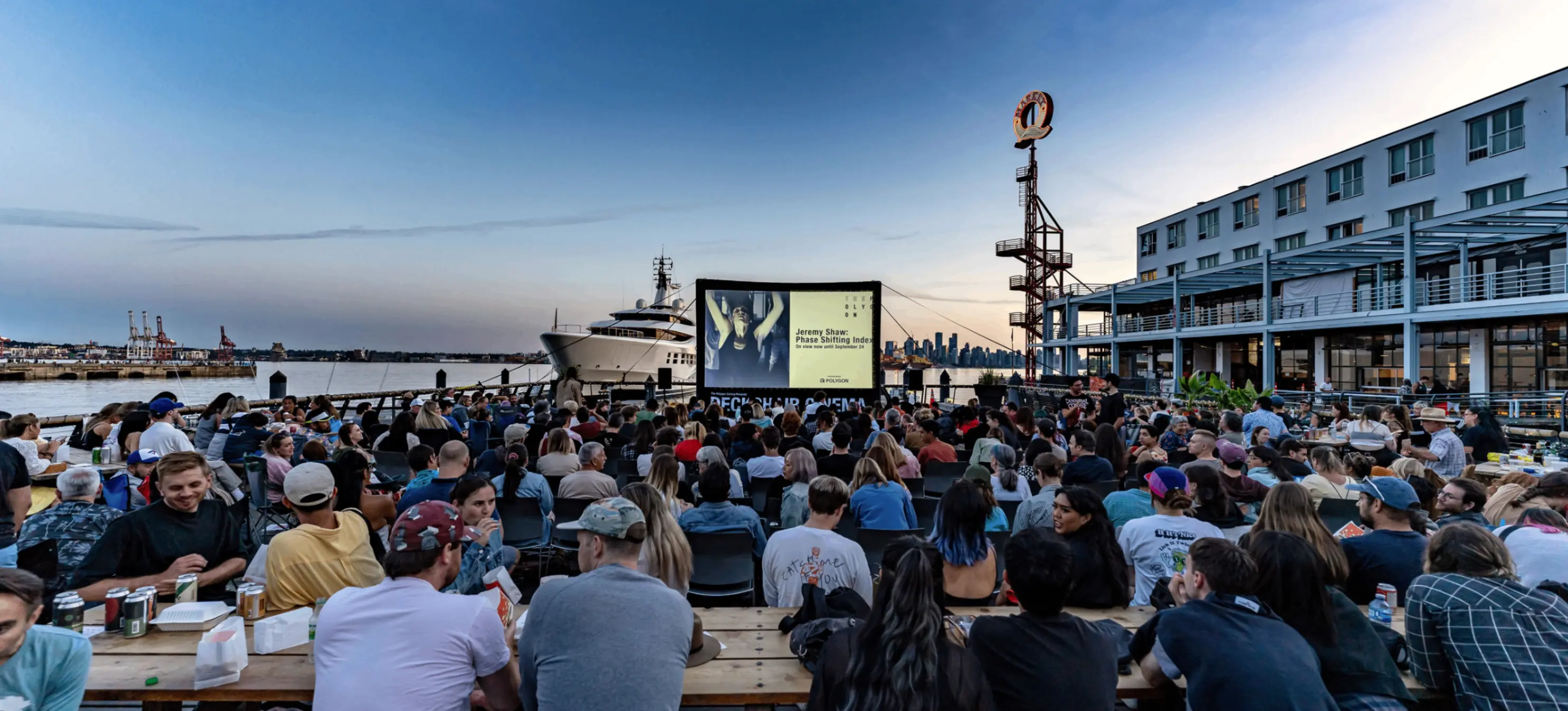 Large crowd seated outdoors on benches watching a movie on a screen near a waterfront with a ship and city skyline in the background at dusk.