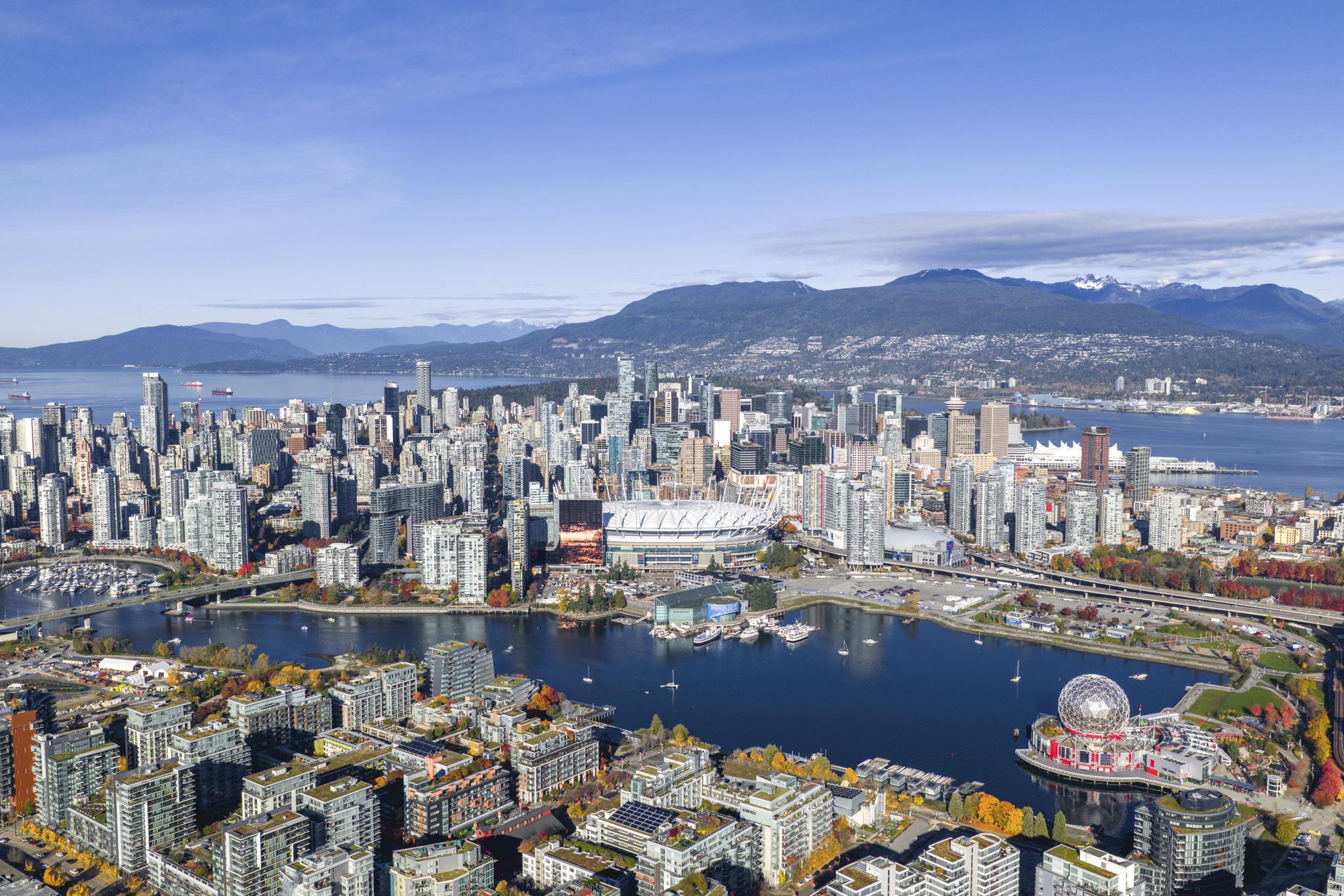 Aerial view of a city skyline with a large stadium, waterfront, and mountains in the background under a clear blue sky.