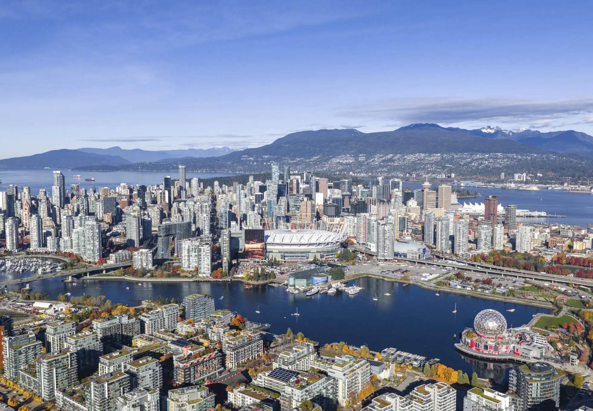 Aerial view of a city skyline with a large stadium, waterfront, and mountains in the background under a clear blue sky.