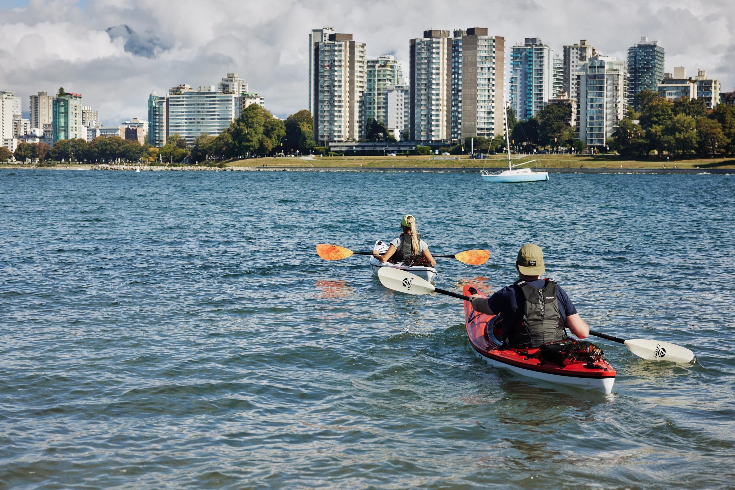 Two people kayaking on a body of water with a city skyline and trees in the background.