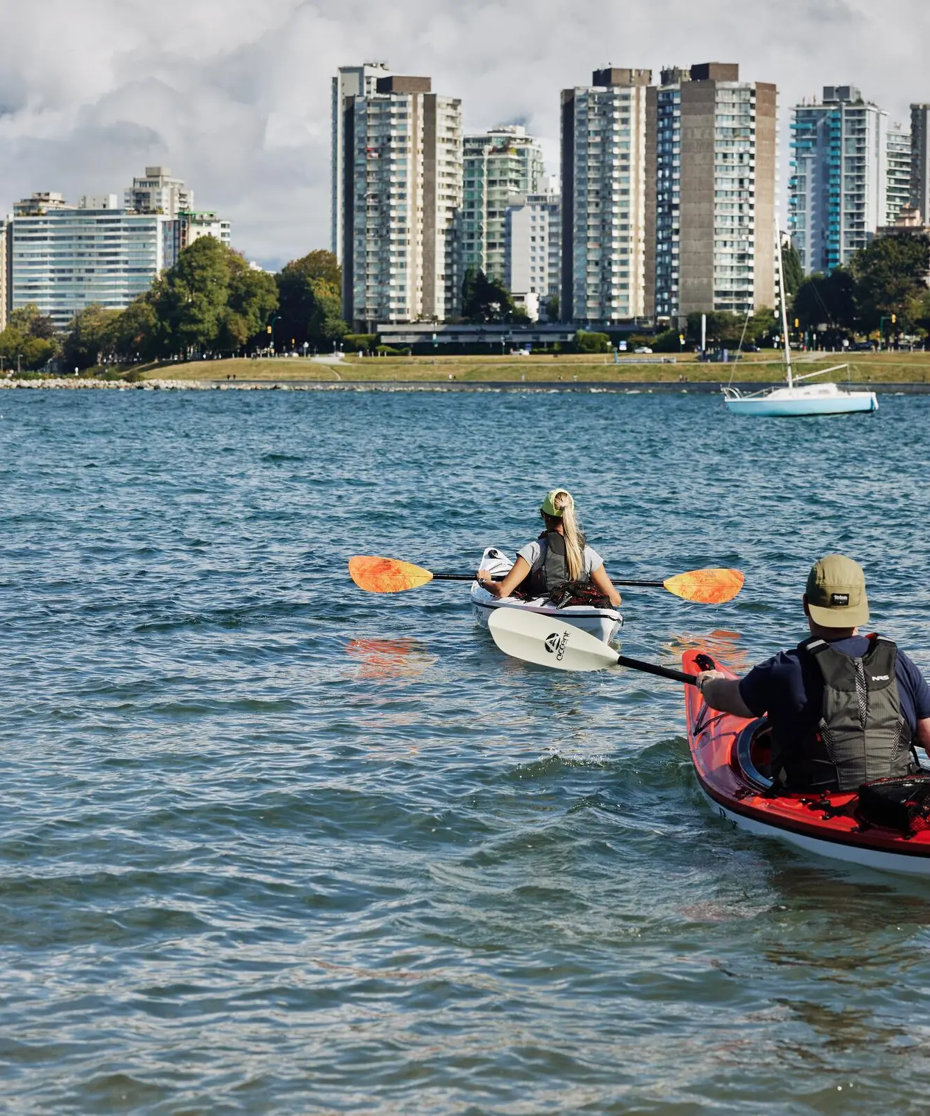 Two people kayaking on a body of water with a city skyline and trees in the background.