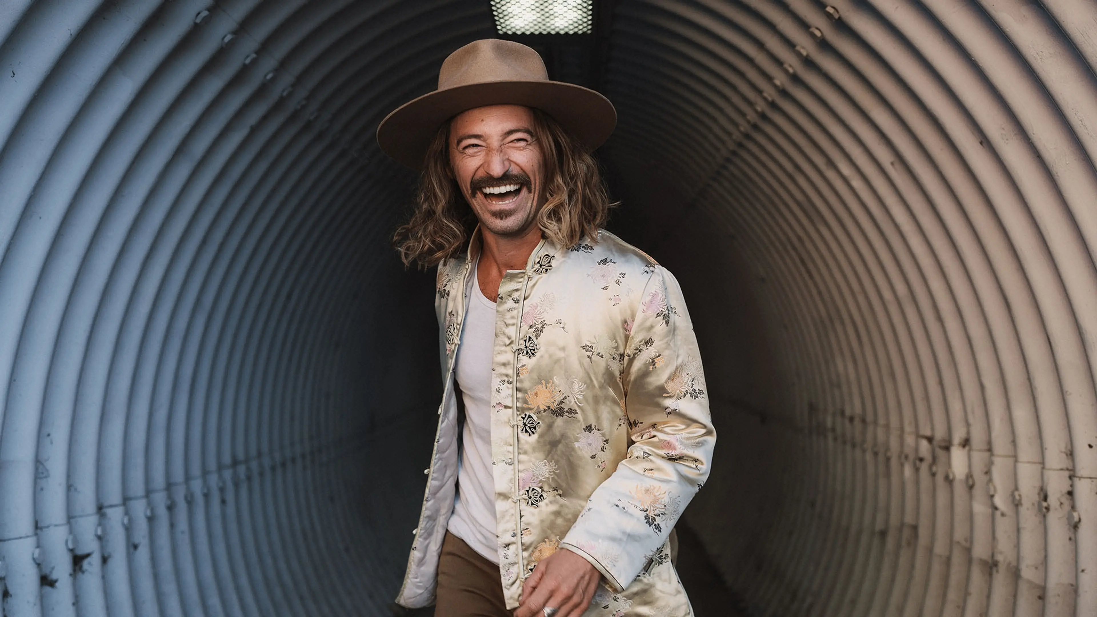 Person with long hair and mustache wearing a wide-brimmed hat and floral jacket, smiling in a corrugated metal tunnel.