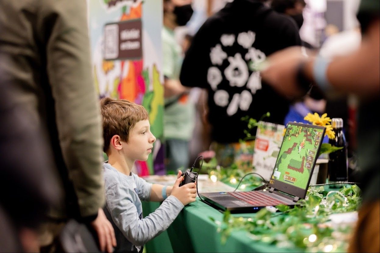 A child holding a game controller playing a pixelated video game on a laptop at a decorated table with plants and lights.