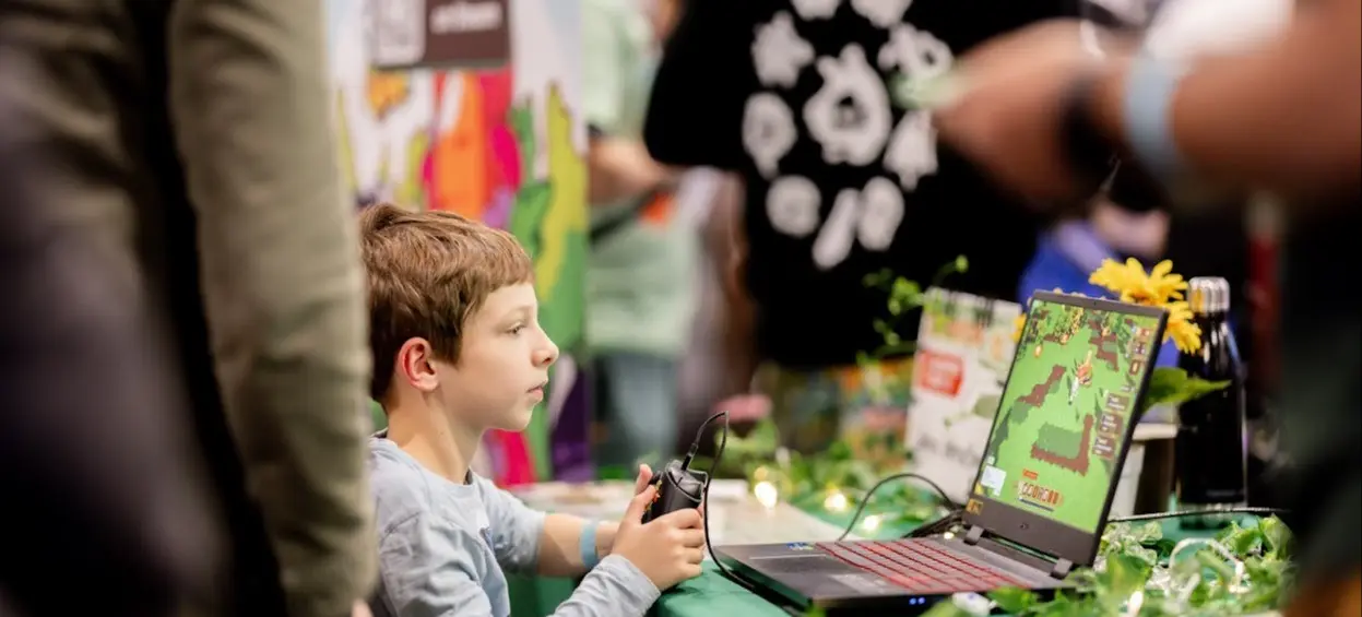 A child holding a game controller playing a pixelated video game on a laptop at a decorated table with plants and lights.