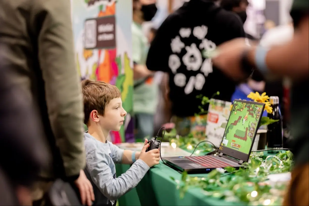 A child holding a game controller playing a pixelated video game on a laptop at a decorated table with plants and lights.