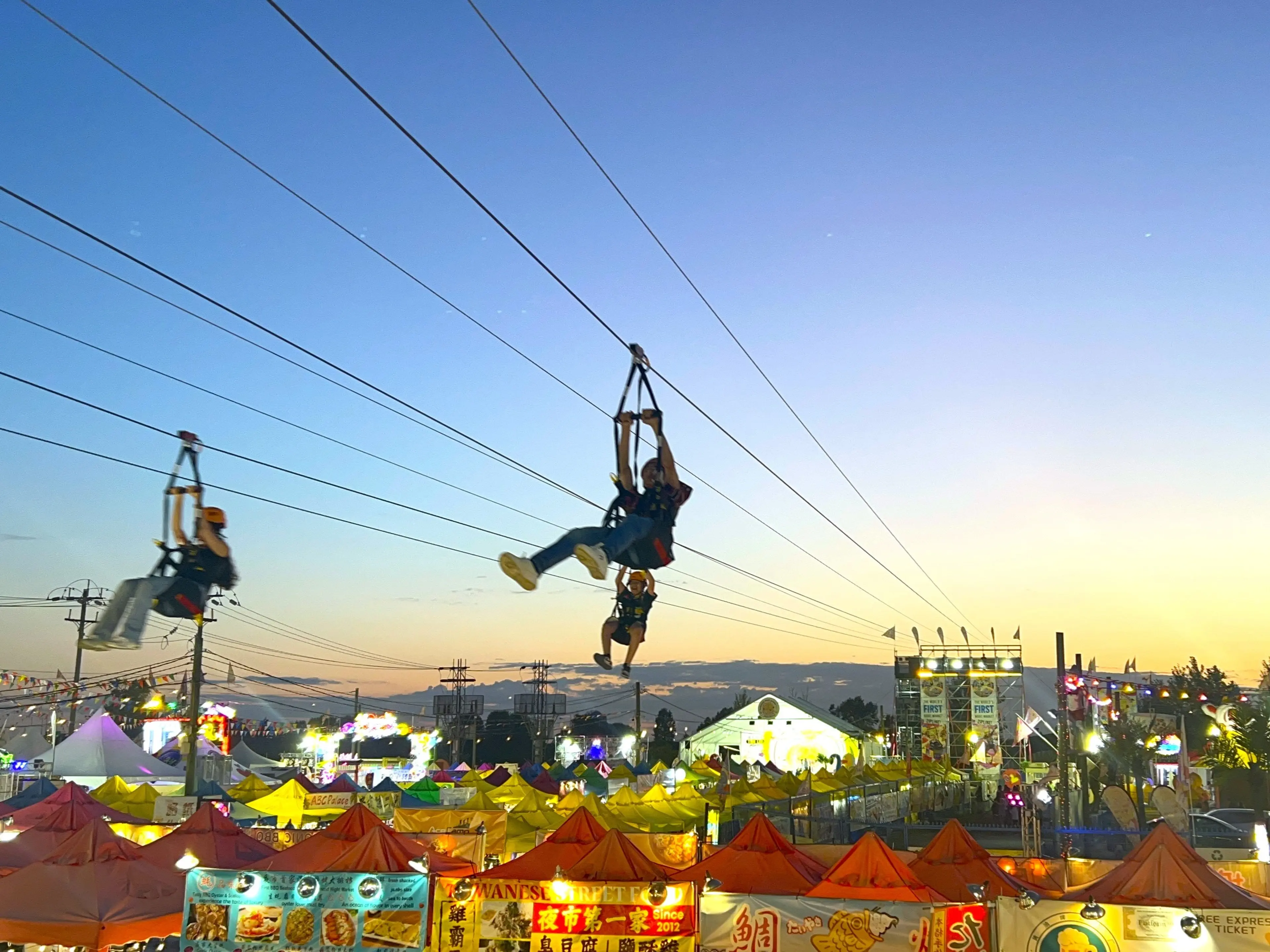 Three people ziplining over a colorful outdoor market at sunset with tents and lights below.