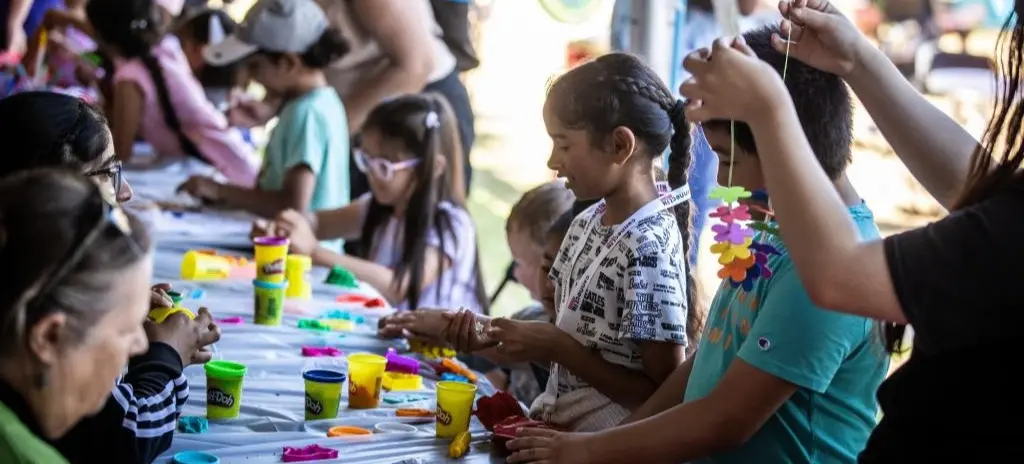 Children and adults sitting at a long table making crafts with colorful Play-Doh and flower decorations.