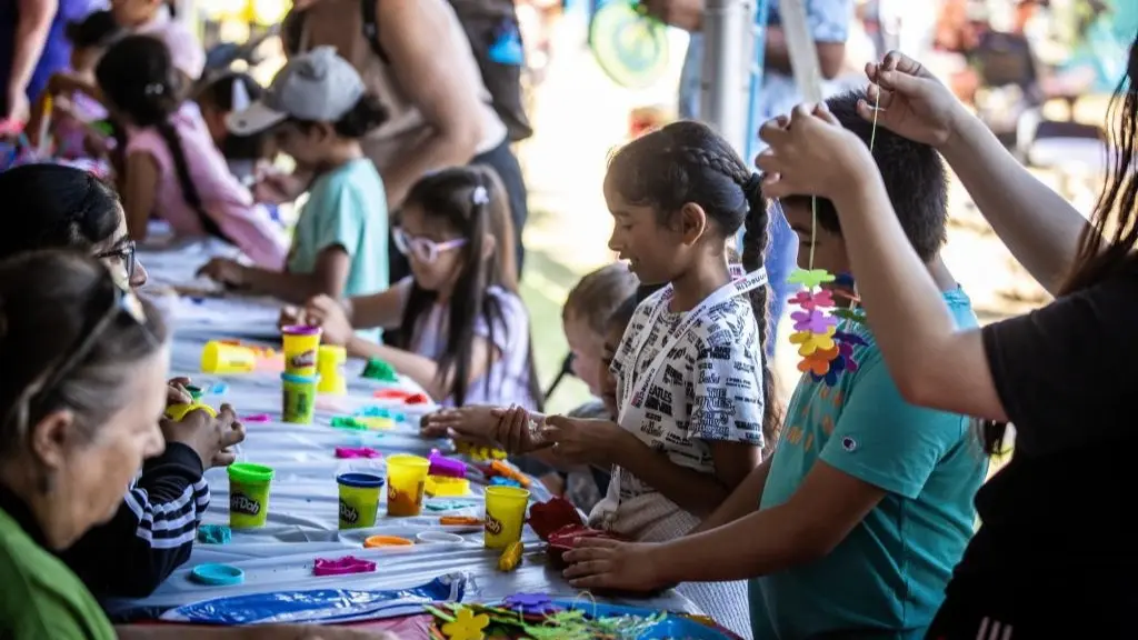 Children and adults sitting at a long table making crafts with colorful Play-Doh and flower decorations.