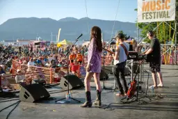 A band performs on an outdoor stage at a music festival, facing a seated crowd. Mountains and blue sky are in the background, with festival banners showcasing Vancouver culture and the vibrant Vancouver art scene. Musical equipment and guitars are on stage.