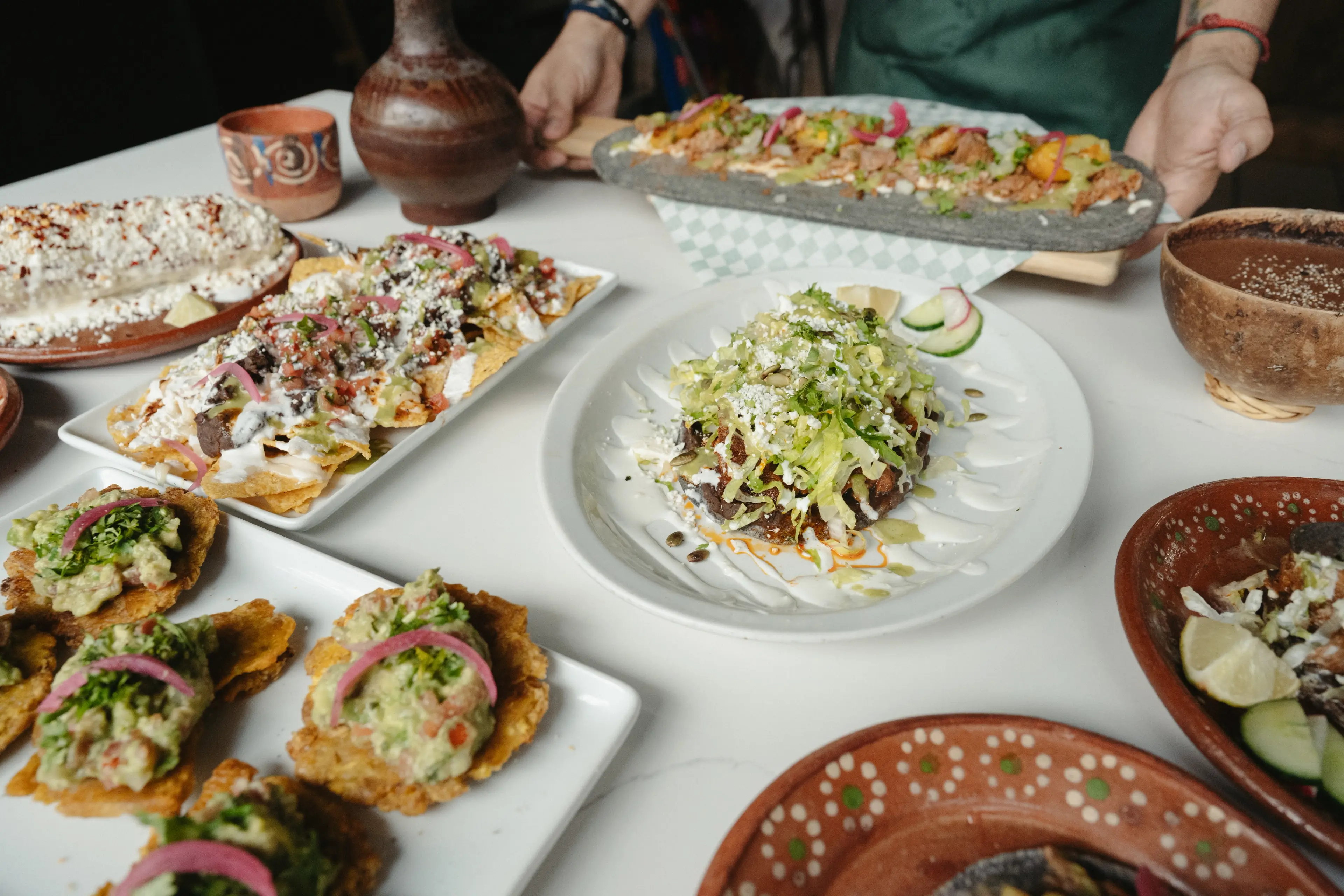 Table with various Mexican dishes including tostadas, nachos, and a person holding a long stone plate with food.