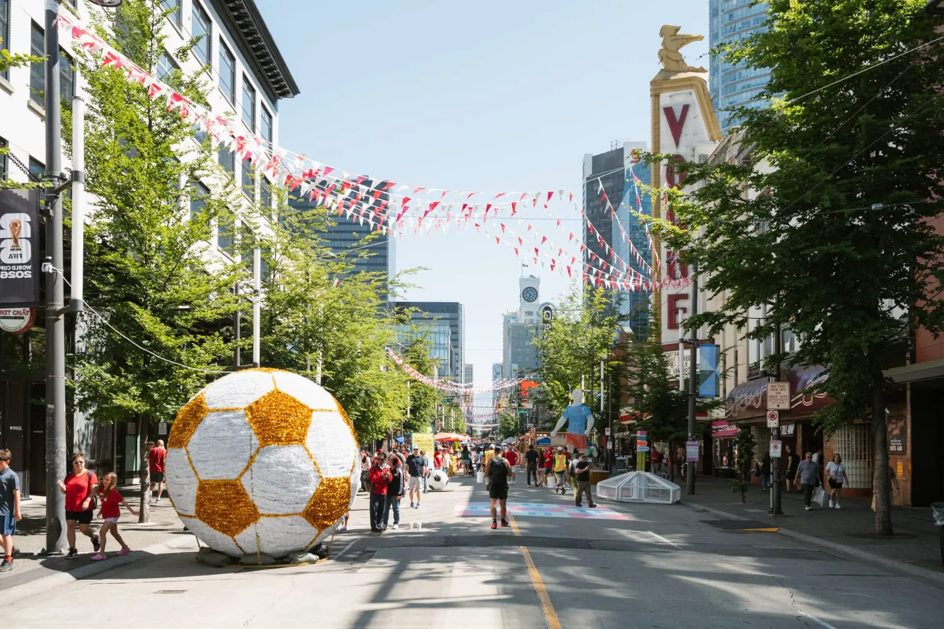 Street decorated with large soccer ball sculptures and red and white flags, people walking and trees lining the road.