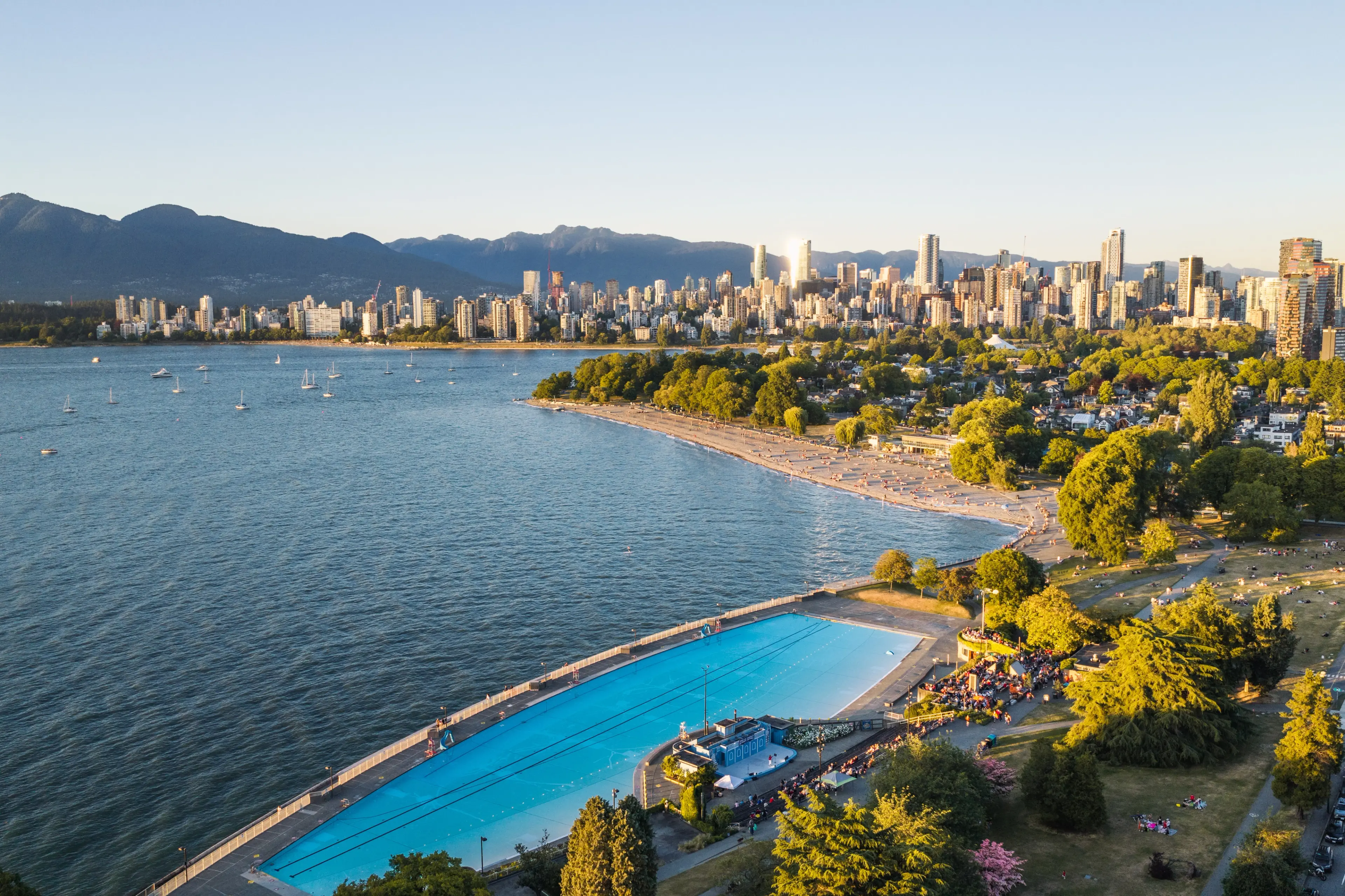 A large outdoor swimming pool near a waterfront park with a city skyline and mountains in the background during sunset.