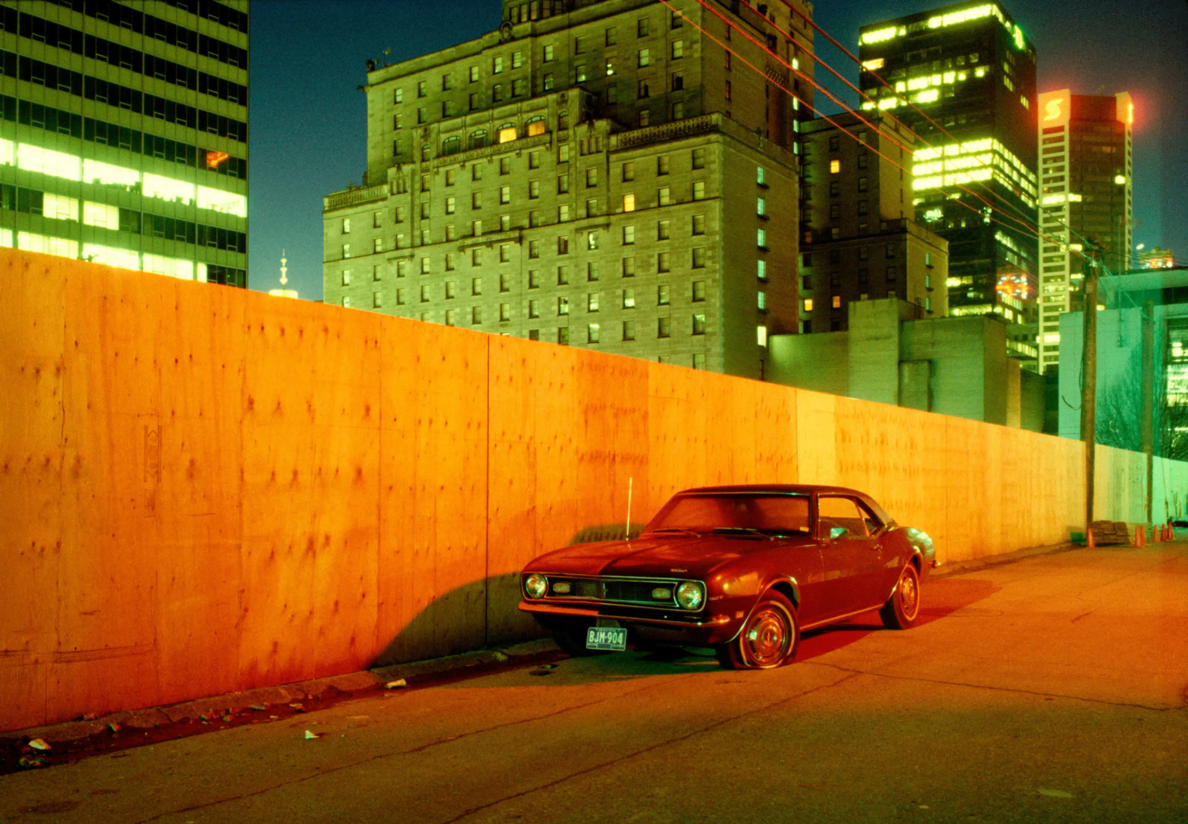 Red vintage car parked on a city street at night with tall buildings and a wooden fence illuminated by yellow-orange light.