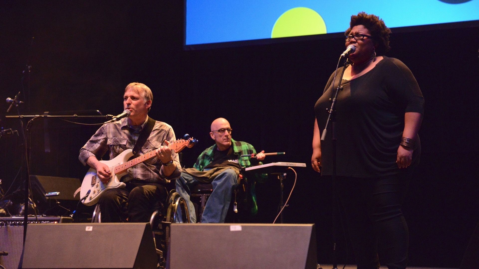 Three people performing on stage: one playing electric guitar and singing, one in wheelchair playing keyboard, and one singing into microphone.