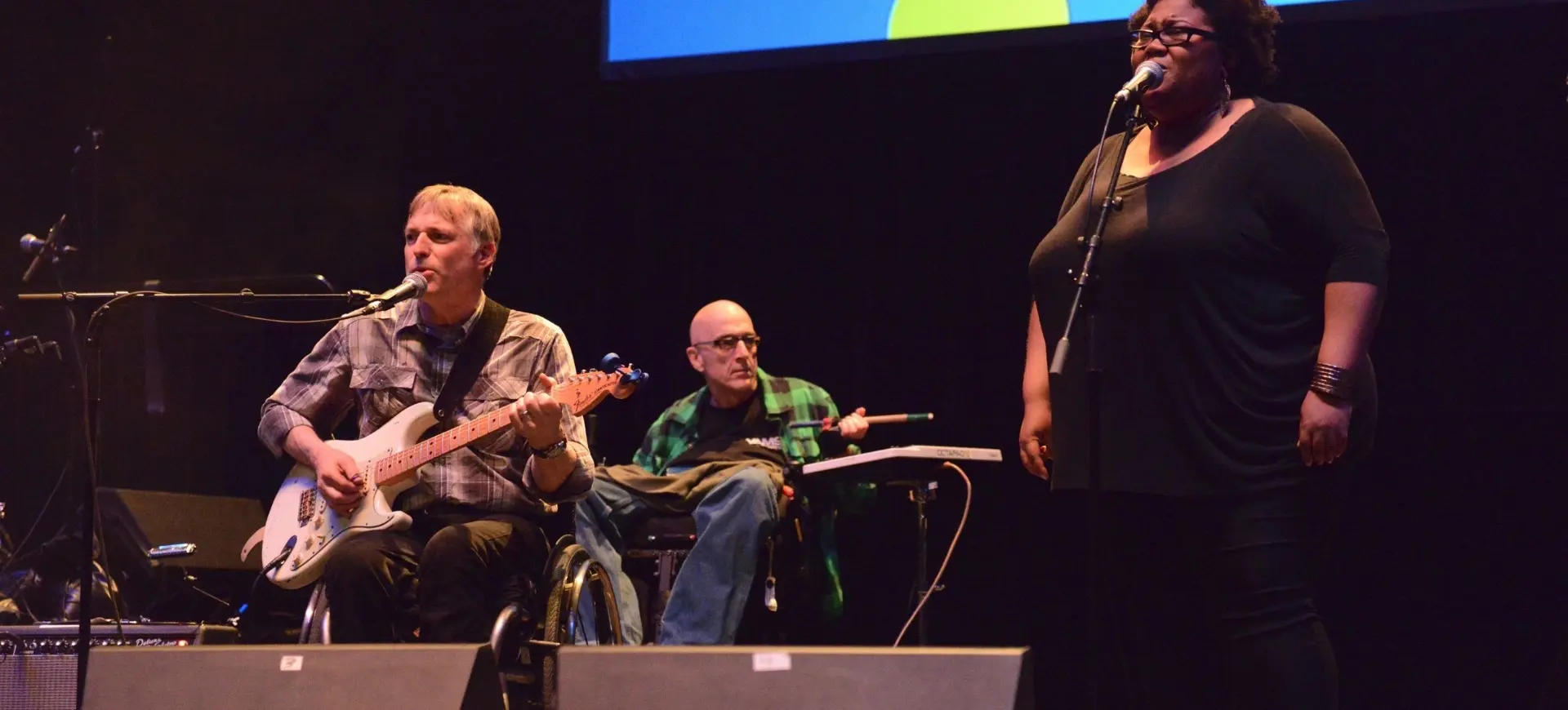Three people performing on stage: one playing electric guitar and singing, one in wheelchair playing keyboard, and one singing into microphone.