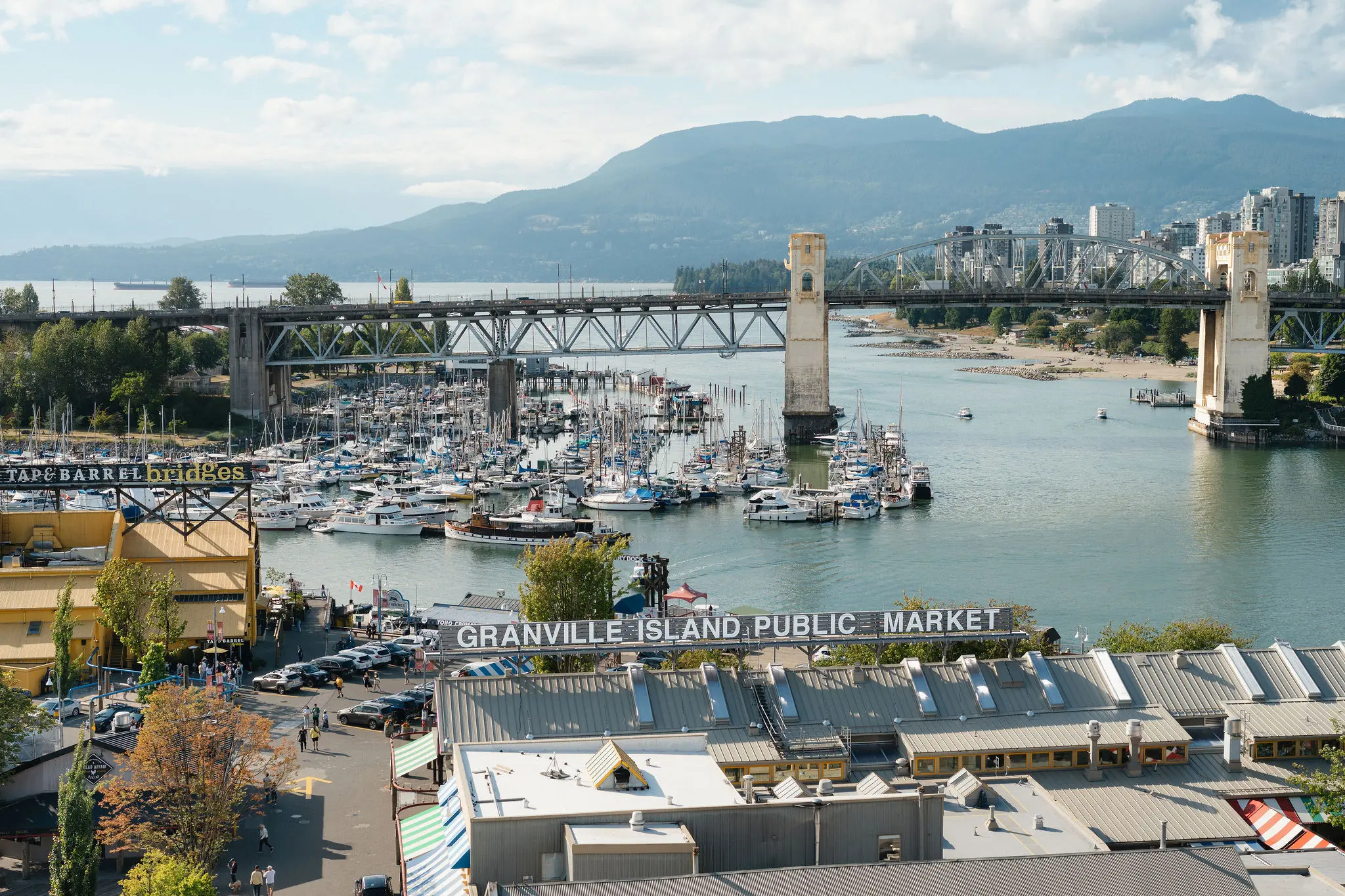 View of Granville Island Public Market, marina with boats, and two bridges over water with mountains in the background.