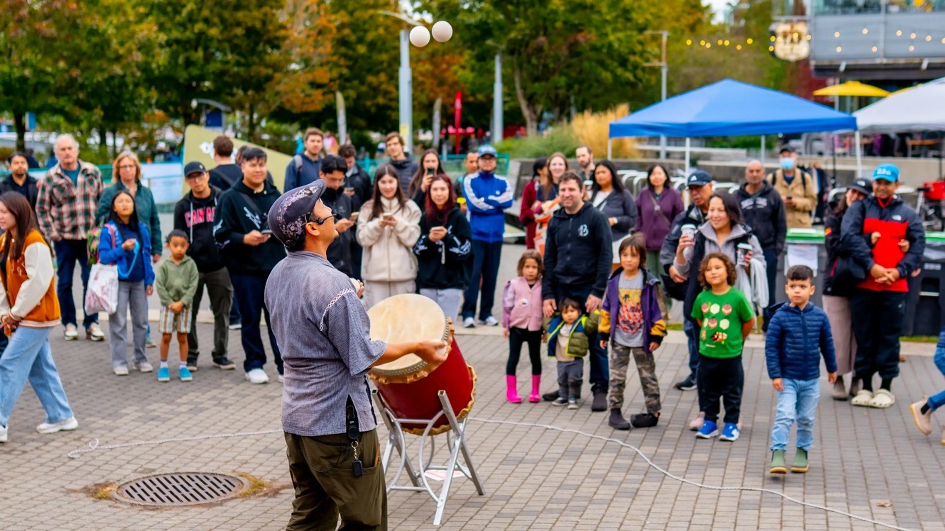A person juggling two balls while holding a drum, with a crowd of people watching outdoors on a paved area.