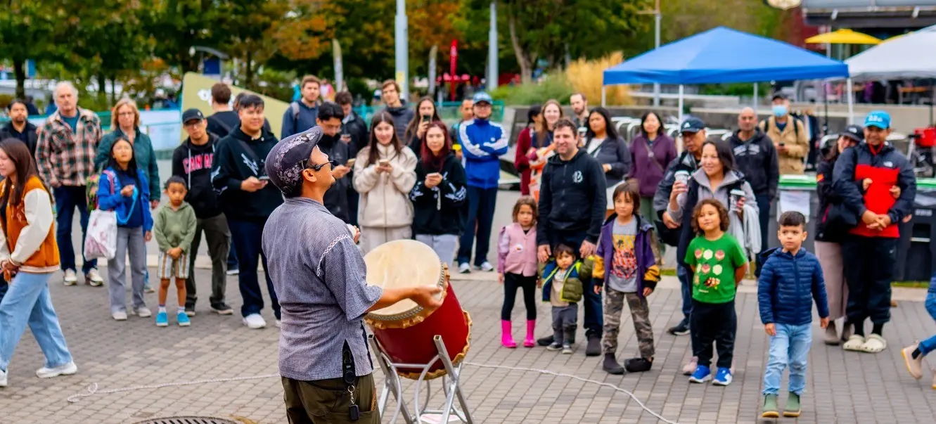 A person juggling two balls while holding a drum, with a crowd of people watching outdoors on a paved area.