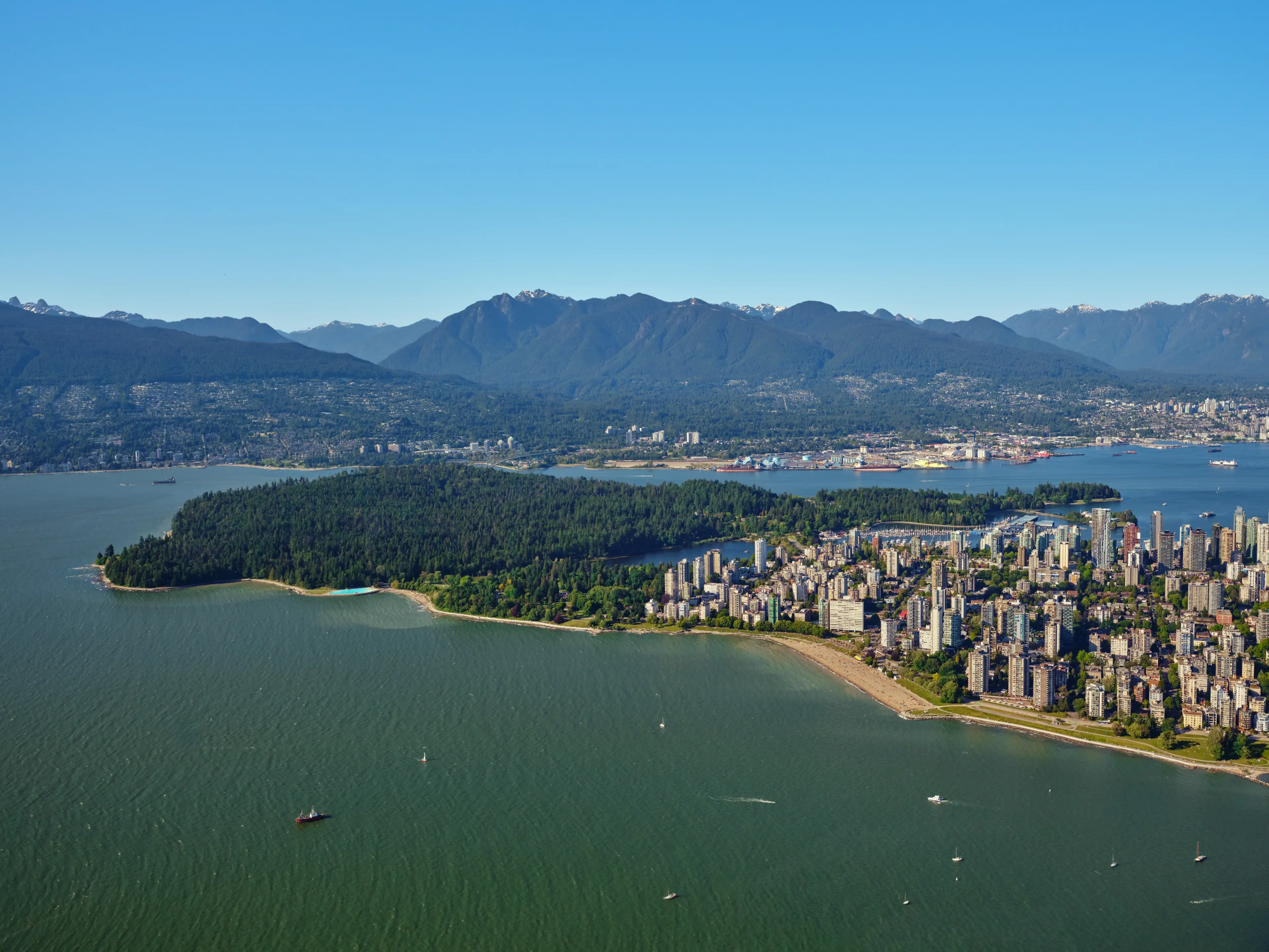 Aerial view of a coastal city with a large forested park, waterfront buildings, and mountains in the background under a clear blue sky.